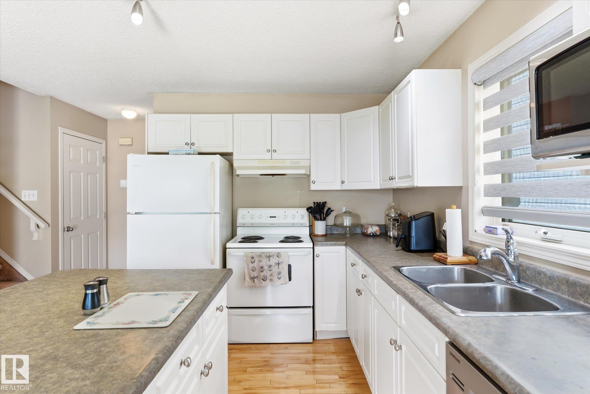 Kitchen featuring white cabinetry, a dual basin stainless steel sink, wood-finish flooring, a kitchen island with a dark countertop, and a window with horizontal blinds - #37 4020 21 St Nw, Edmonton, AB - Indoor Photo Showing Kitchen With Double Sink