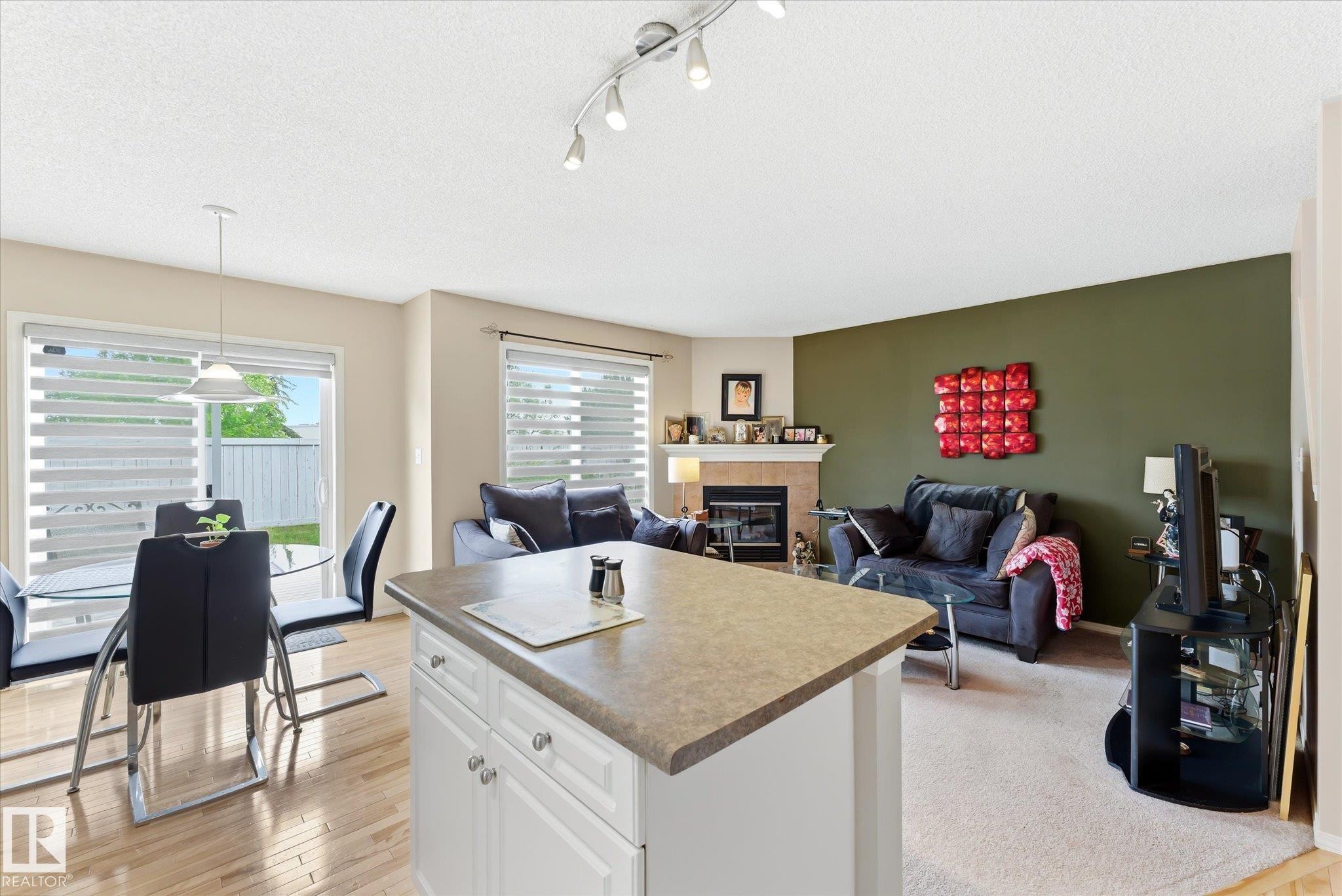Open-concept living space featuring wood-finish flooring, a kitchen island with storage, a dining area with a glass patio door, a living room with carpet flooring, and a fireplace with a mantel - #37 4020 21 St Nw, Edmonton, AB - Indoor With Fireplace