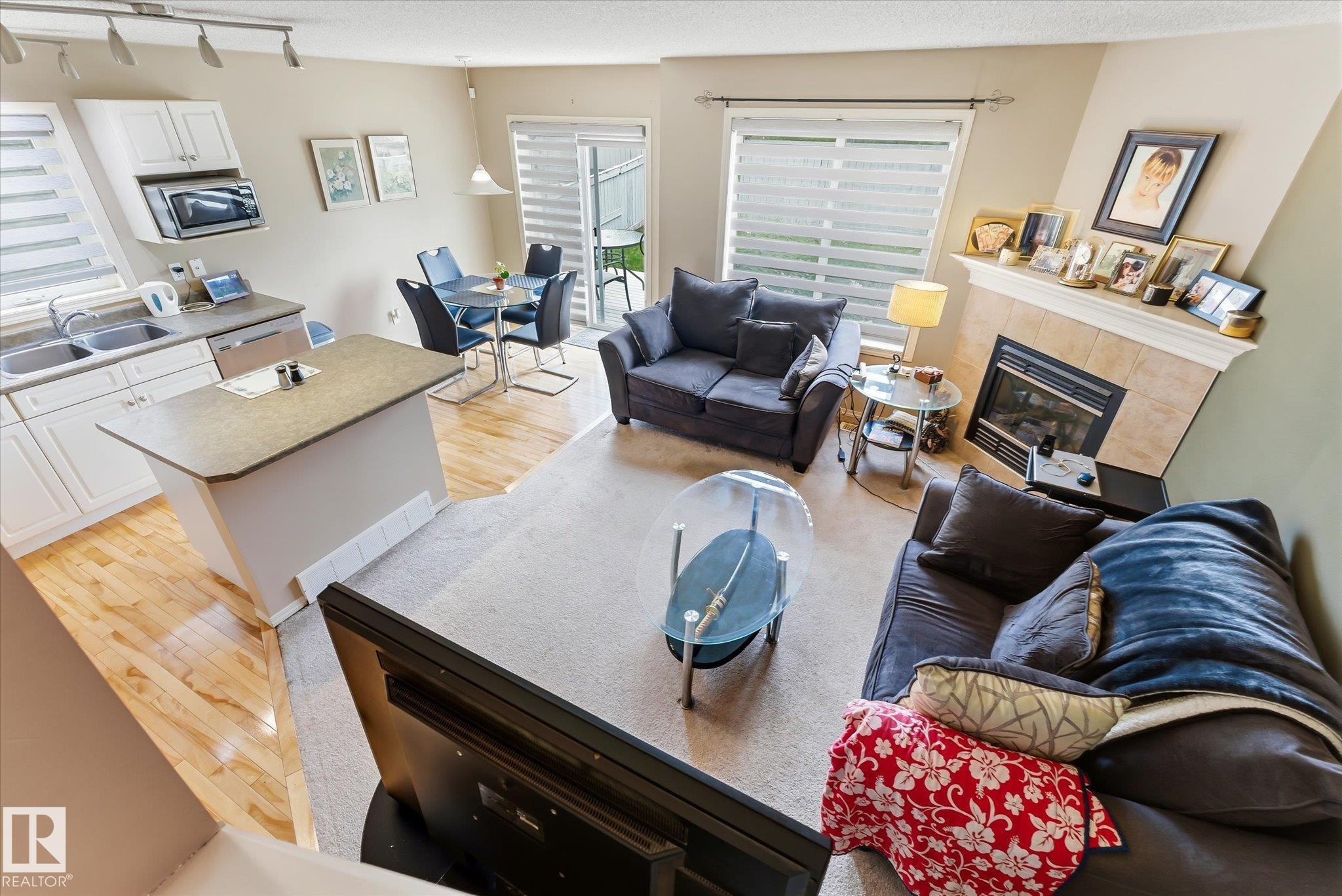 Open-concept living area featuring a kitchen island, wood-finish flooring, a dining area with direct outdoor access, a fireplace with a tiled surround, and track lighting - #37 4020 21 St Nw, Edmonton, AB - Indoor Photo Showing Living Room With Fireplace