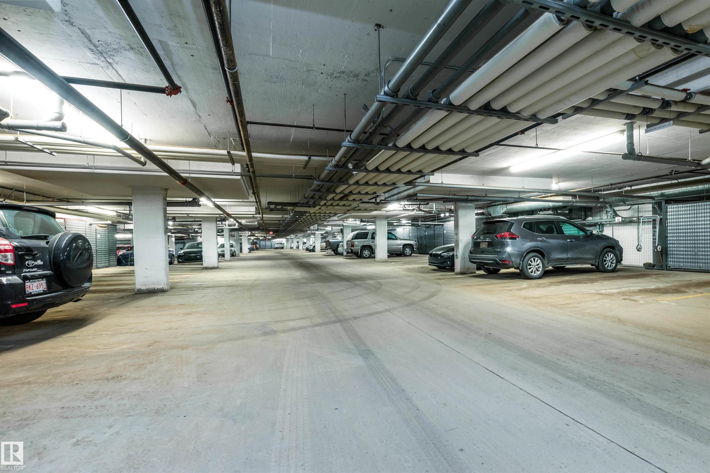 Underground parking garage featuring concrete flooring, structural support columns, overhead fluorescent lighting, exposed utility piping, and individual storage units - 439 300 Palisades Way, Sherwood Park, AB - Indoor Photo Showing Garage