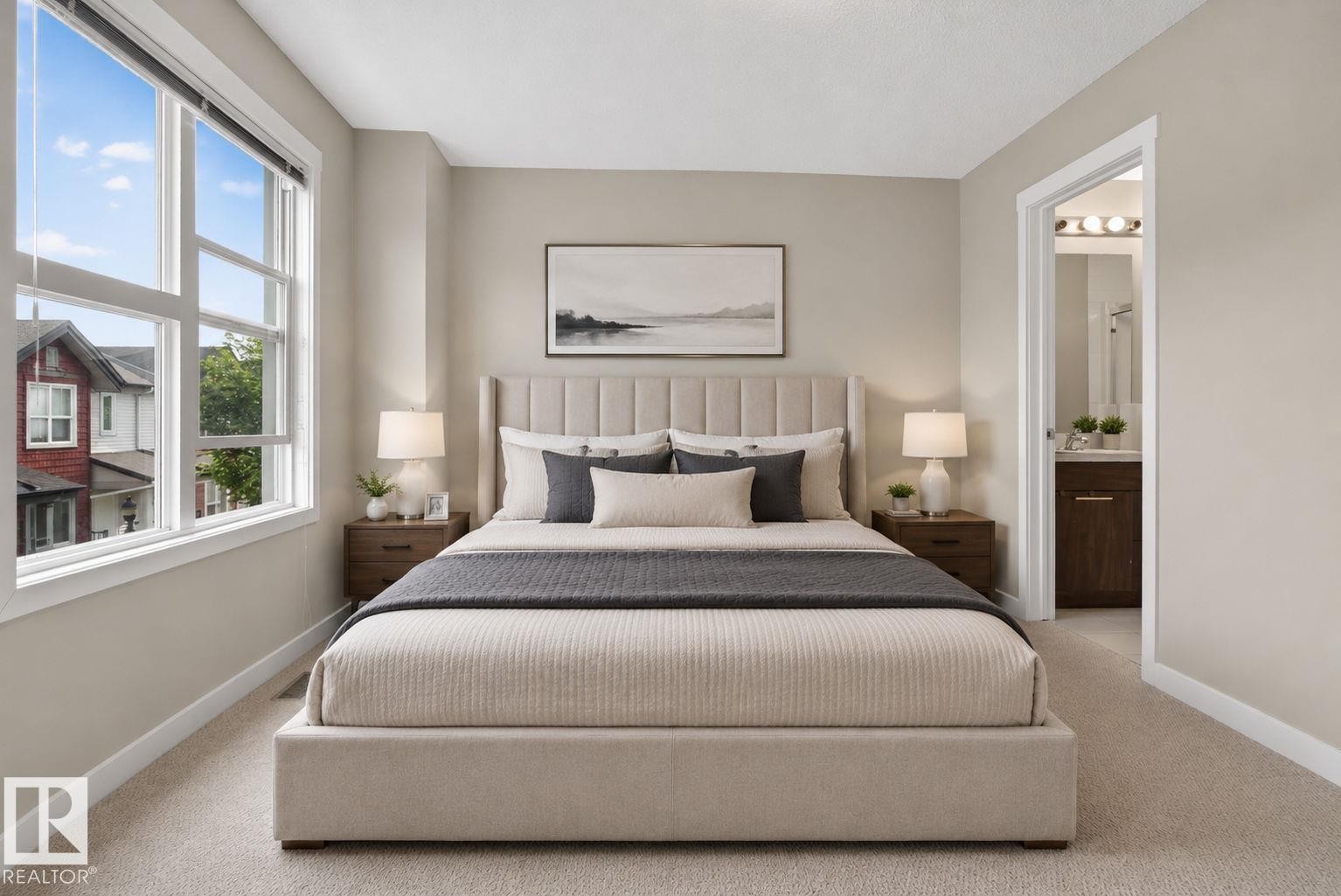 Spacious bedroom featuring light neutral wall tones, plush carpet flooring, large multi-pane windows, and a doorway opening to an en-suite bathroom with a dark wood-finish vanity - 30 6075 Schonsee Way, Edmonton, AB