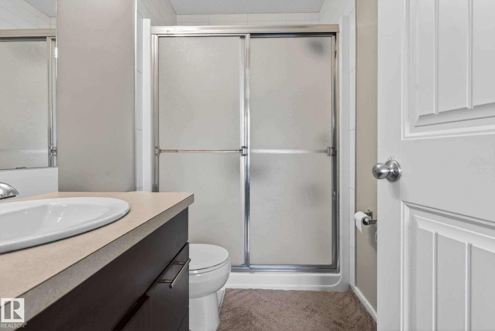 Bathroom featuring a sliding glass shower enclosure, a dark wood-finish vanity with an integrated basin, a light-toned countertop, and a white paneled door with chrome hardware - 30 6075 Schonsee Way, Edmonton, AB