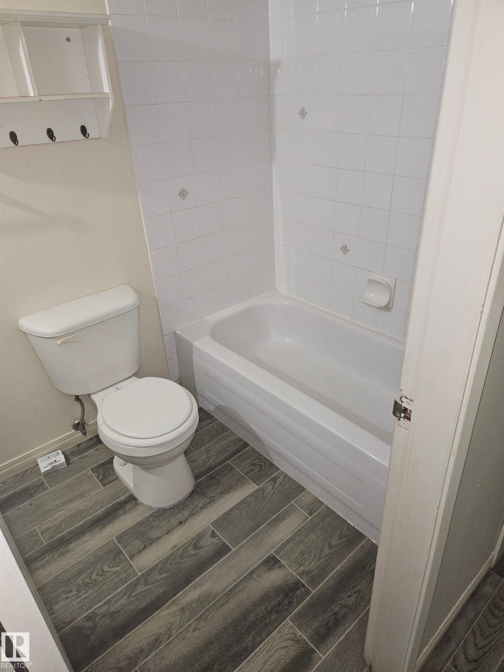 Bathroom featuring a white toilet, white bathtub with white tiled surround, and wood-finish tile flooring - 401 10615 110 Street, Edmonton, AB