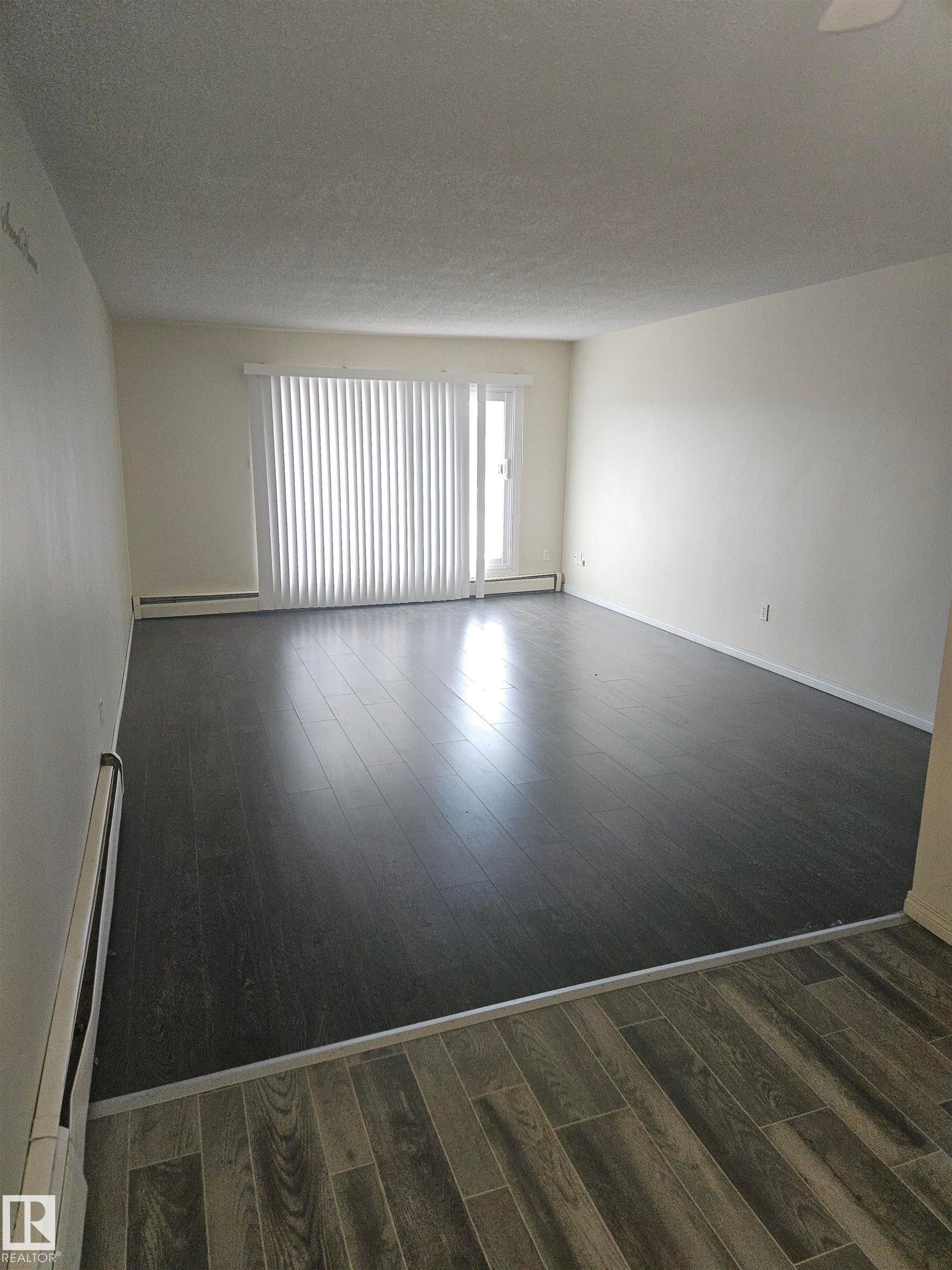 Spacious room featuring dark wood-finish flooring, light-colored walls, and a sliding glass door with vertical blinds - 401 10615 110 Street, Edmonton, AB