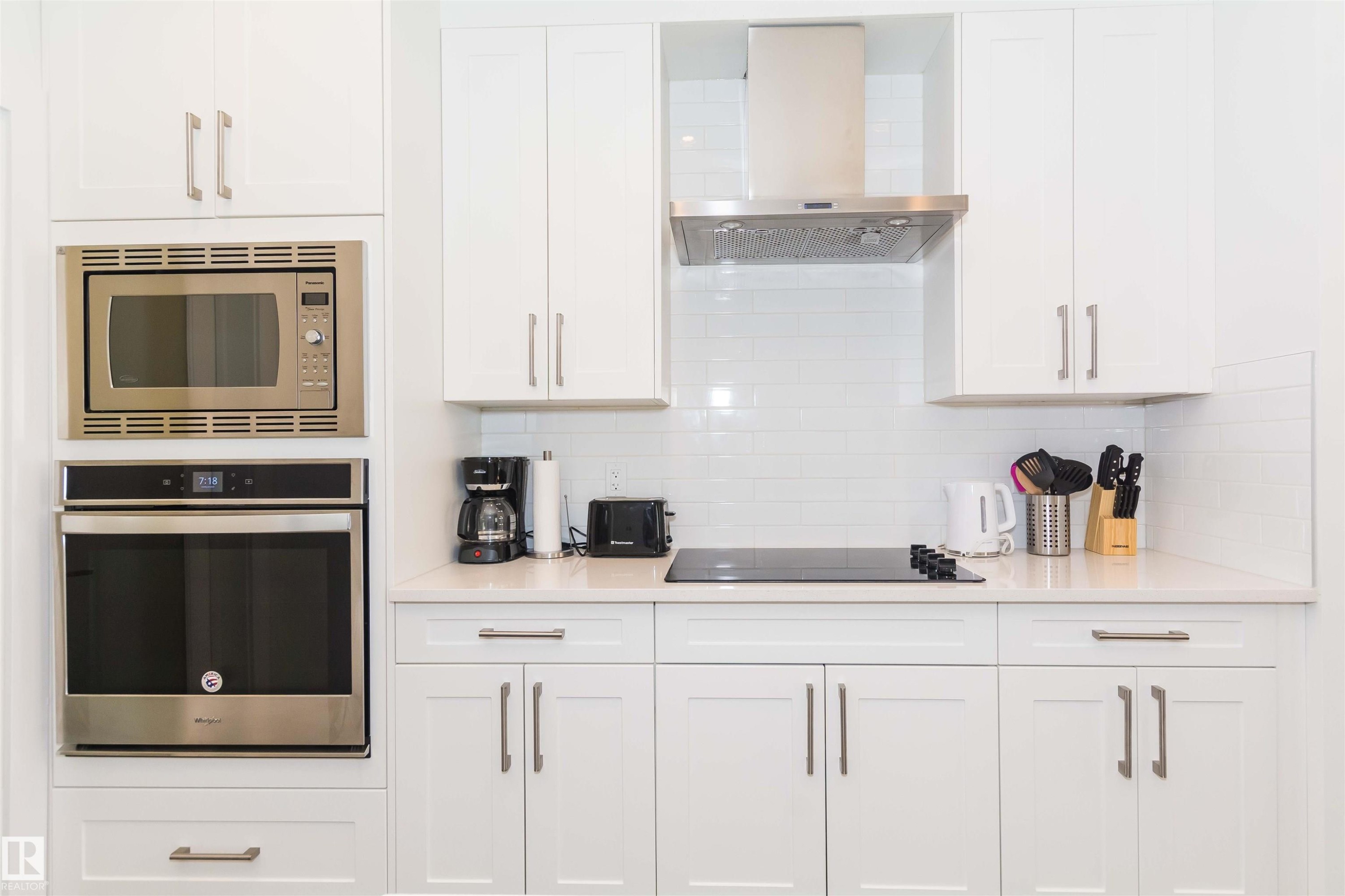 Modern kitchen featuring white shaker cabinetry, stainless steel built-in microwave and oven, a sleek cooktop, and a matching stainless steel range hood - 15168 16 Street Nw, Edmonton, AB