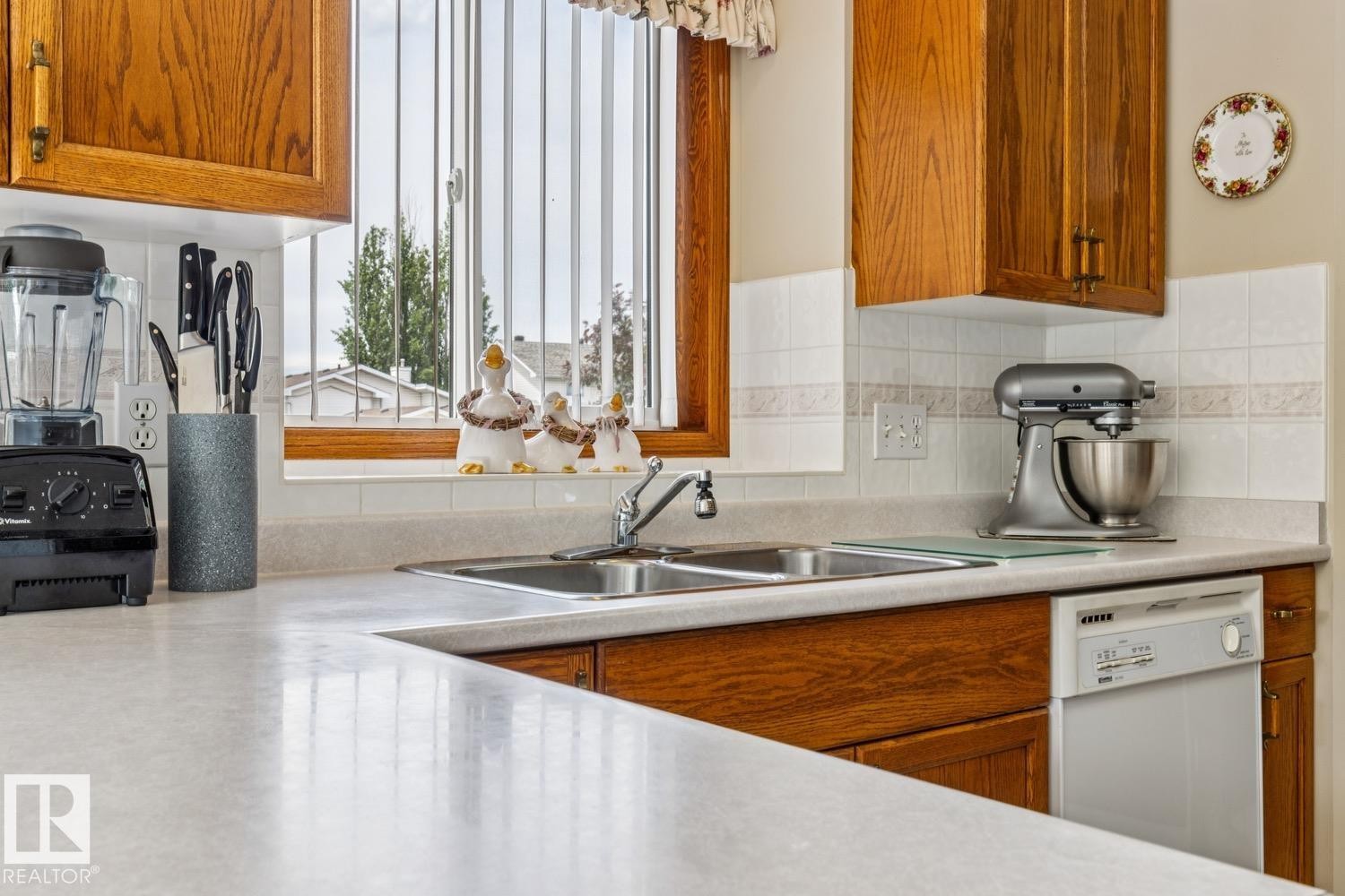 Kitchen featuring a double basin stainless steel sink, light-toned countertops, wood cabinetry, a tiled backsplash, and a built-in dishwasher - 5131 152A Avenue, Edmonton, AB