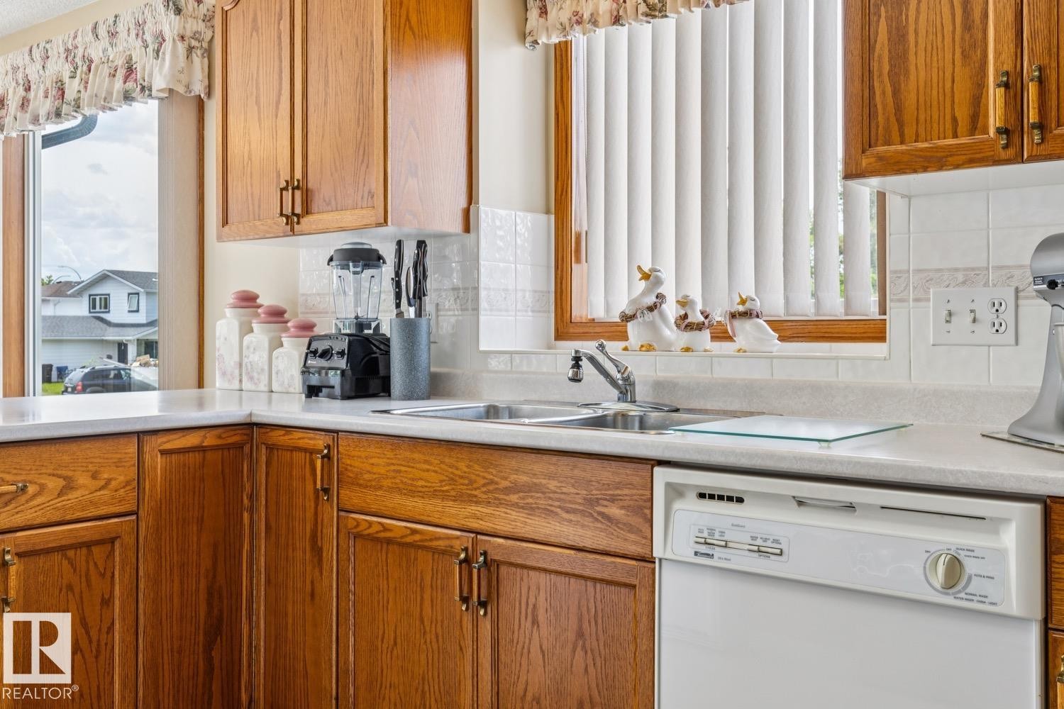 Kitchen featuring wood cabinetry with bronze hardware, a double basin stainless steel sink, and a white built-in dishwasher - 5131 152A Avenue, Edmonton, AB