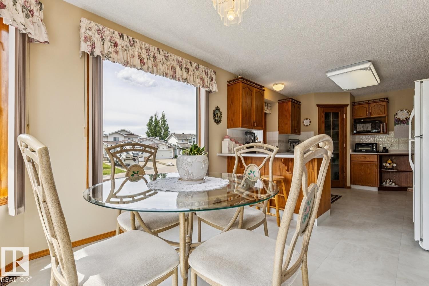 Dining area with a rounded glass-top table, light-toned upholstered chairs, and large bay windows - 5131 152A Avenue, Edmonton, AB