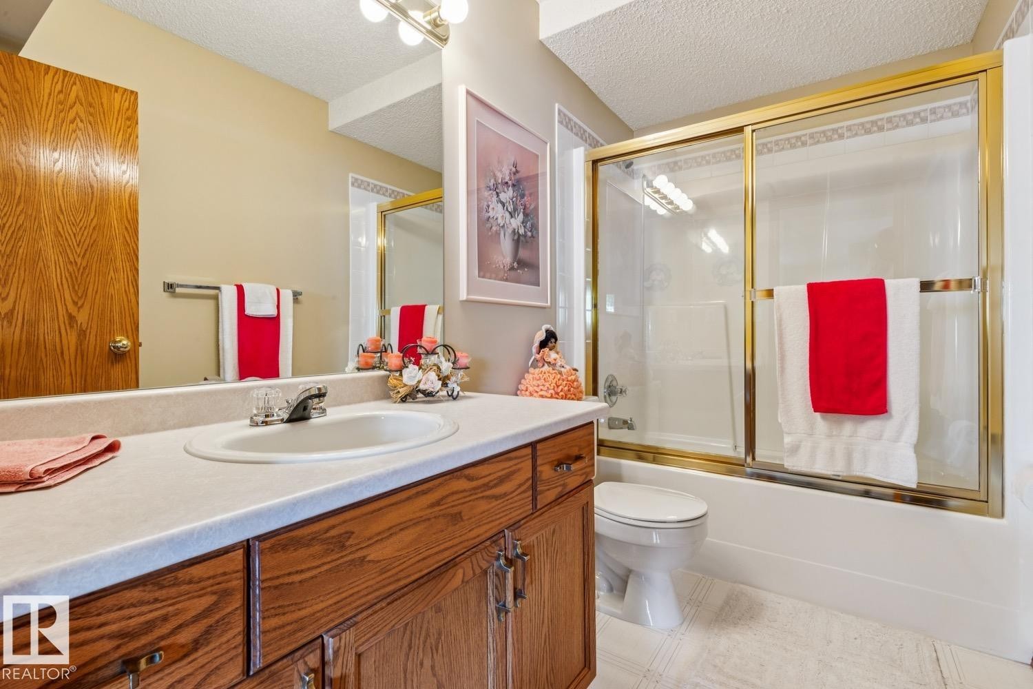 Bathroom featuring a vanity with an integrated sink, wood-finish cabinetry, a large mirror, a shower-tub combination with a gold-finish enclosure, and textured ceiling - 5131 152A Avenue, Edmonton, AB