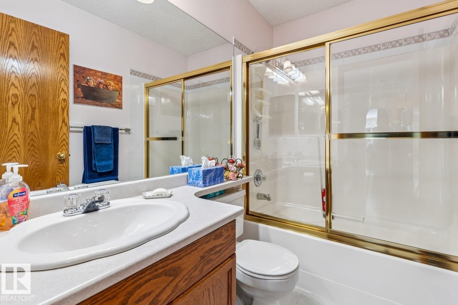 Bathroom featuring a wood-finish vanity with an integrated sink, a large wall-mounted mirror, and a tub with a gold-tone framed glass enclosure - 5131 152A Avenue, Edmonton, AB