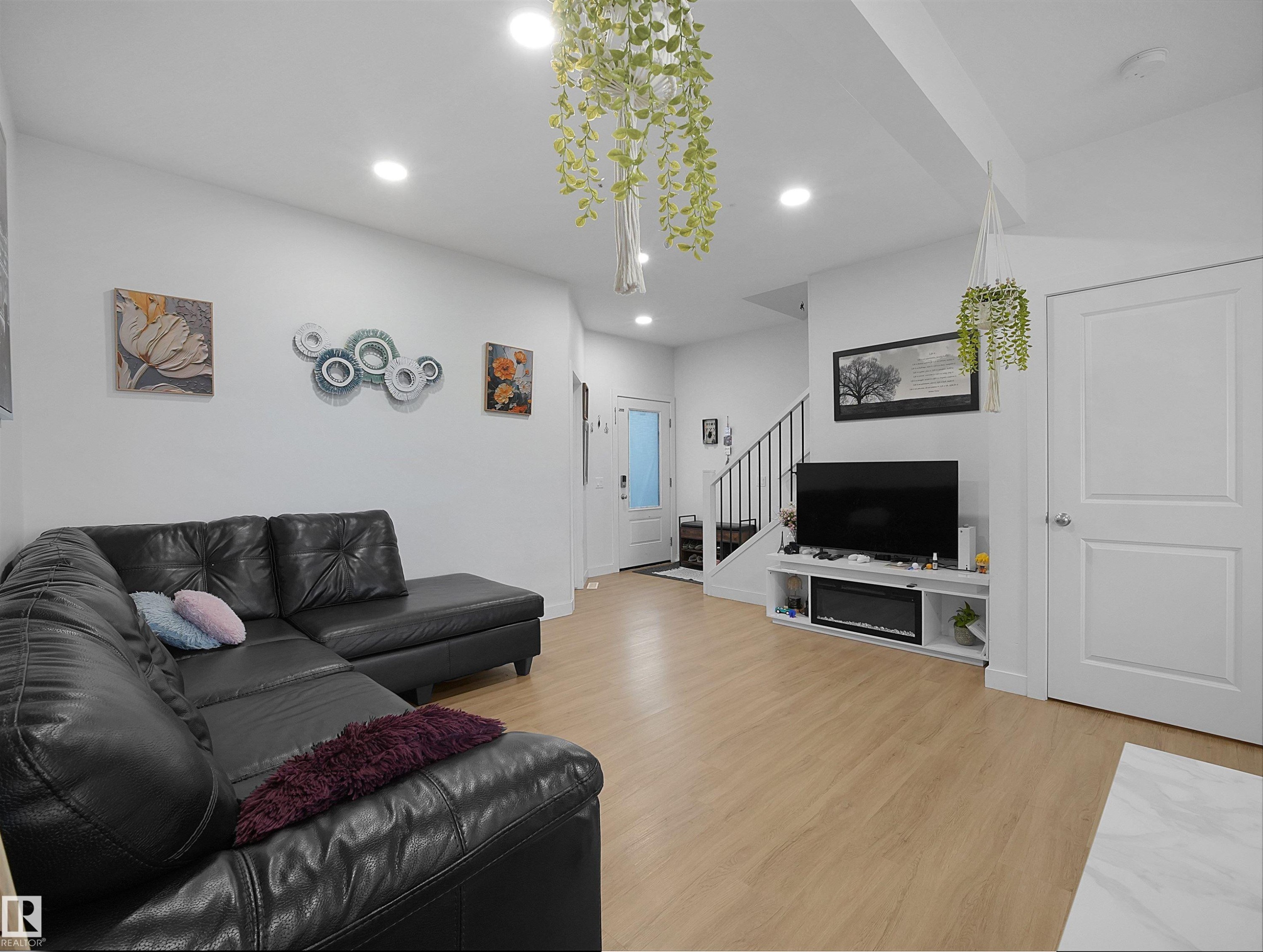 Spacious living area featuring light wood-finish flooring, recessed ceiling lighting, a built-in media console with an electric fireplace, a white paneled door, and a staircase with dark metal railings - 19316 29 Avenue, Edmonton, AB