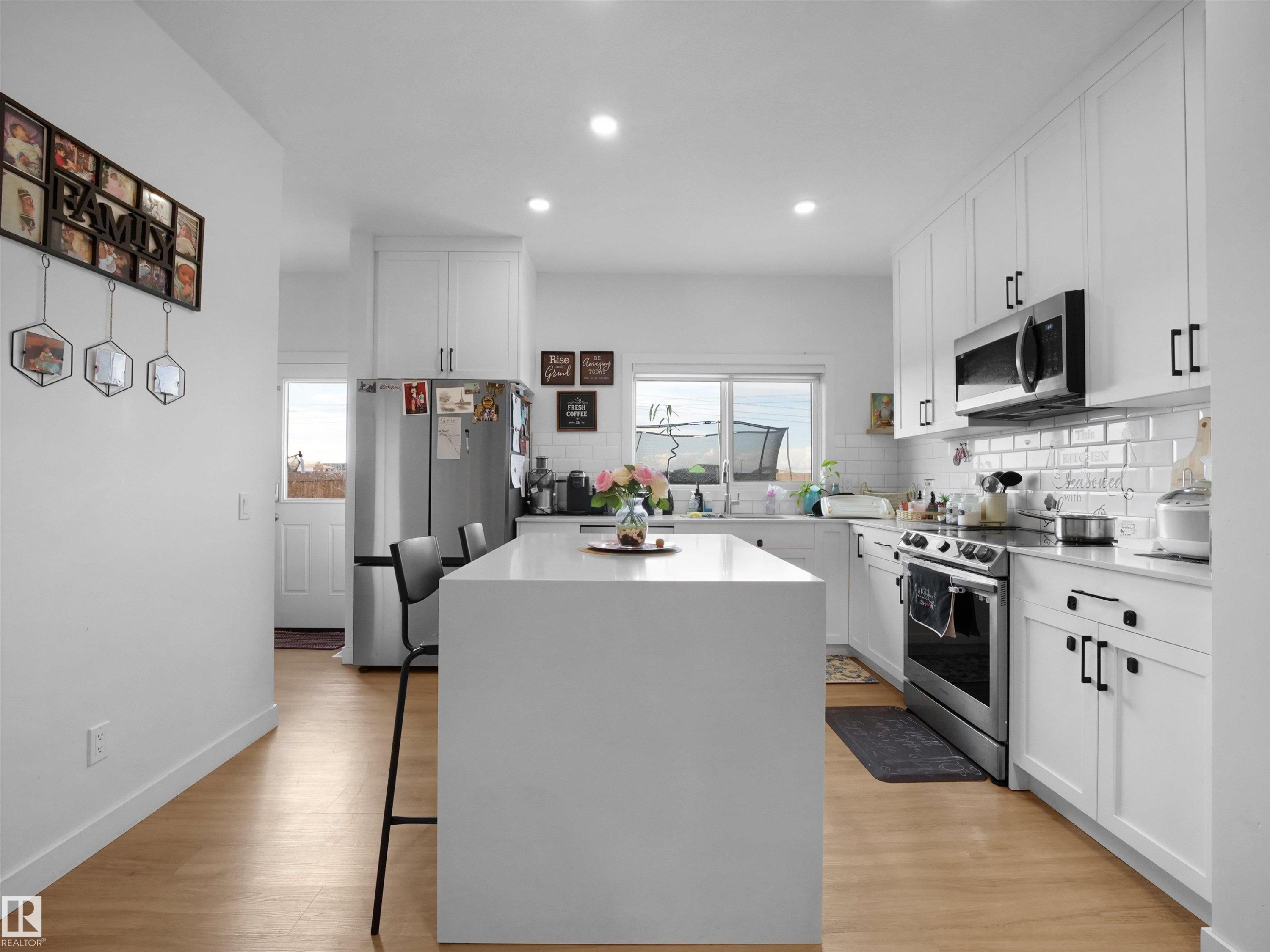 Modern kitchen featuring a large central island with seating, white shaker cabinetry, stainless steel appliances, and wood-finish flooring - 19316 29 Avenue, Edmonton, AB