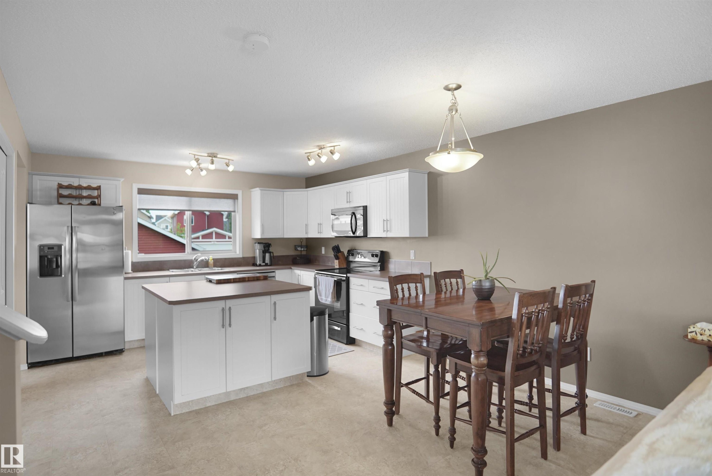 Kitchen with stainless steel appliances, white cabinetry, a central island with a wood-finish countertop, and a dining area featuring a dark wood table with matching chairs - 292 Simmonds Way, Leduc, AB