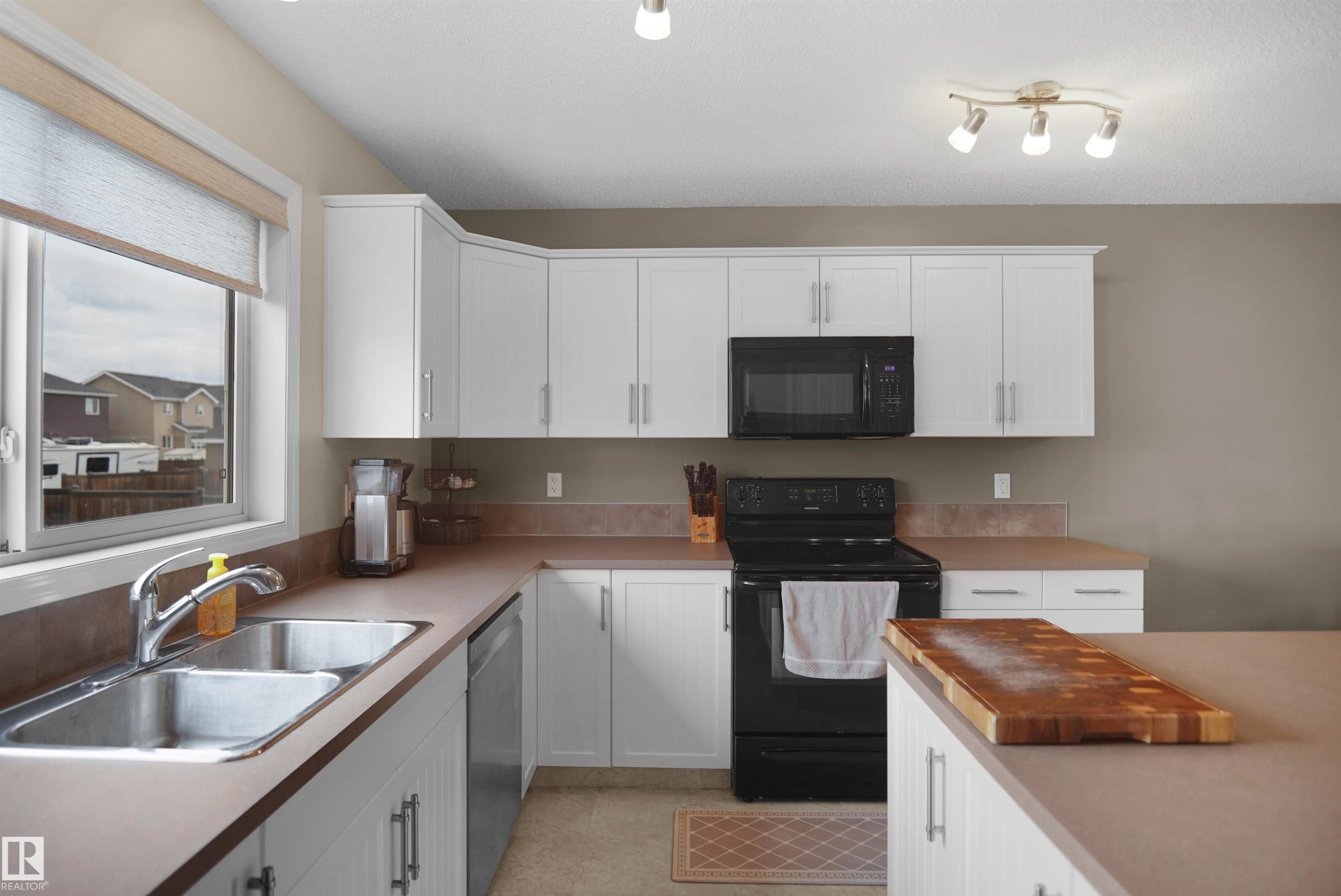 Kitchen featuring white cabinetry, a double basin stainless steel sink, black built-in microwave, black electric range, and light brown countertops - 292 Simmonds Way, Leduc, AB