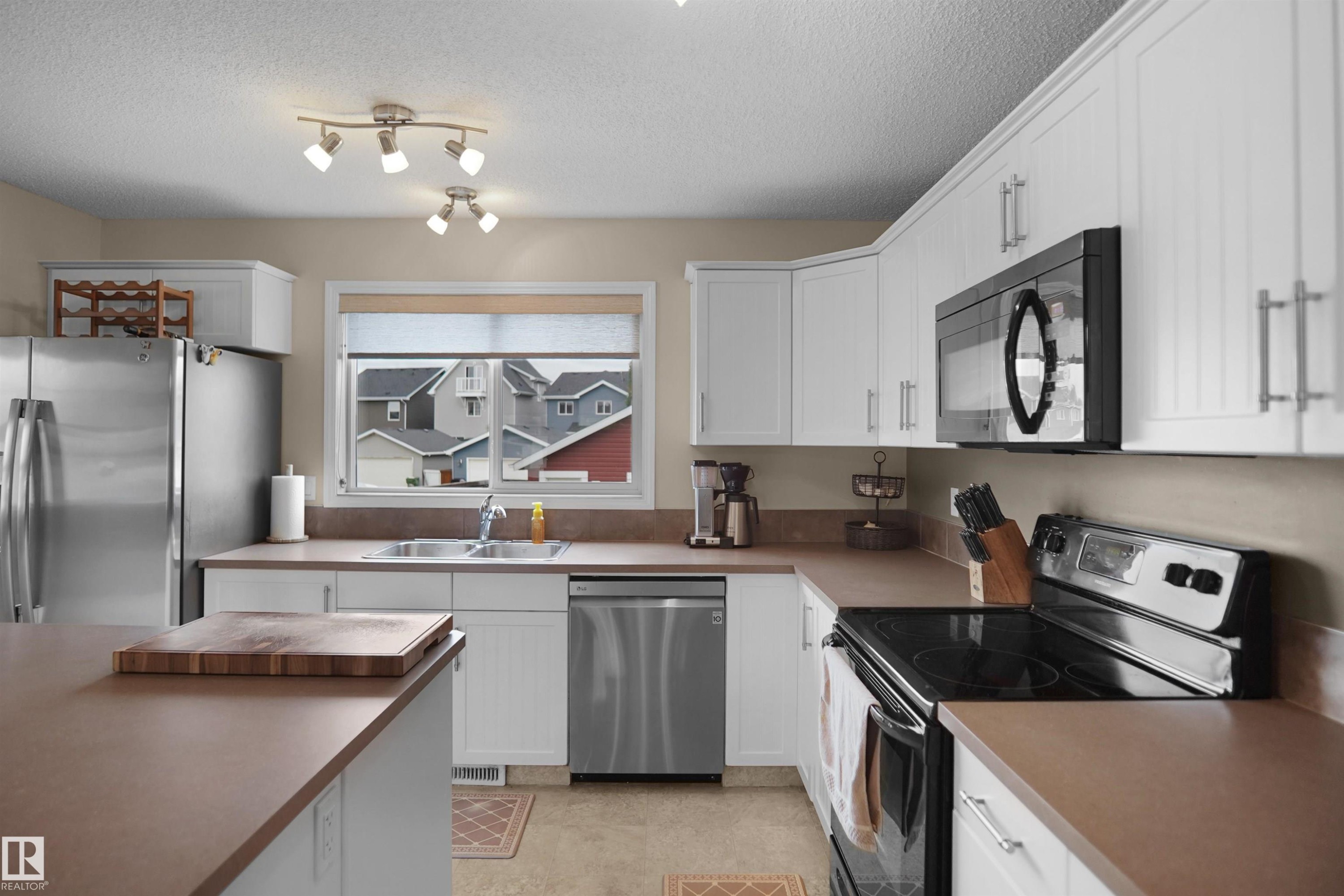 Functional kitchen featuring white cabinetry, stainless steel appliances, track lighting, a double basin sink, and a built-in microwave - 292 Simmonds Way, Leduc, AB