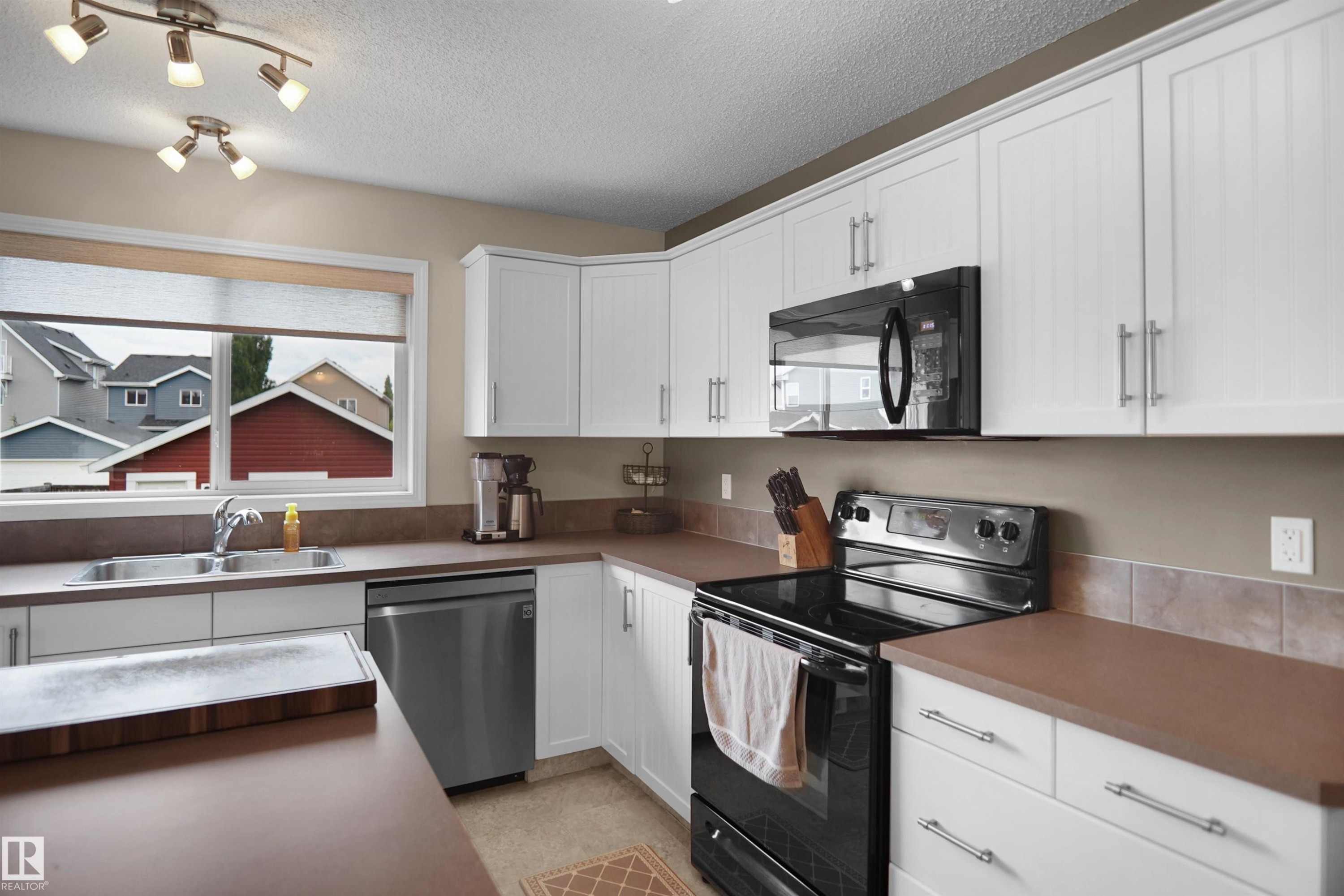 Kitchen featuring white cabinetry, a double basin sink, black appliances, track lighting, and a window - 292 Simmonds Way, Leduc, AB