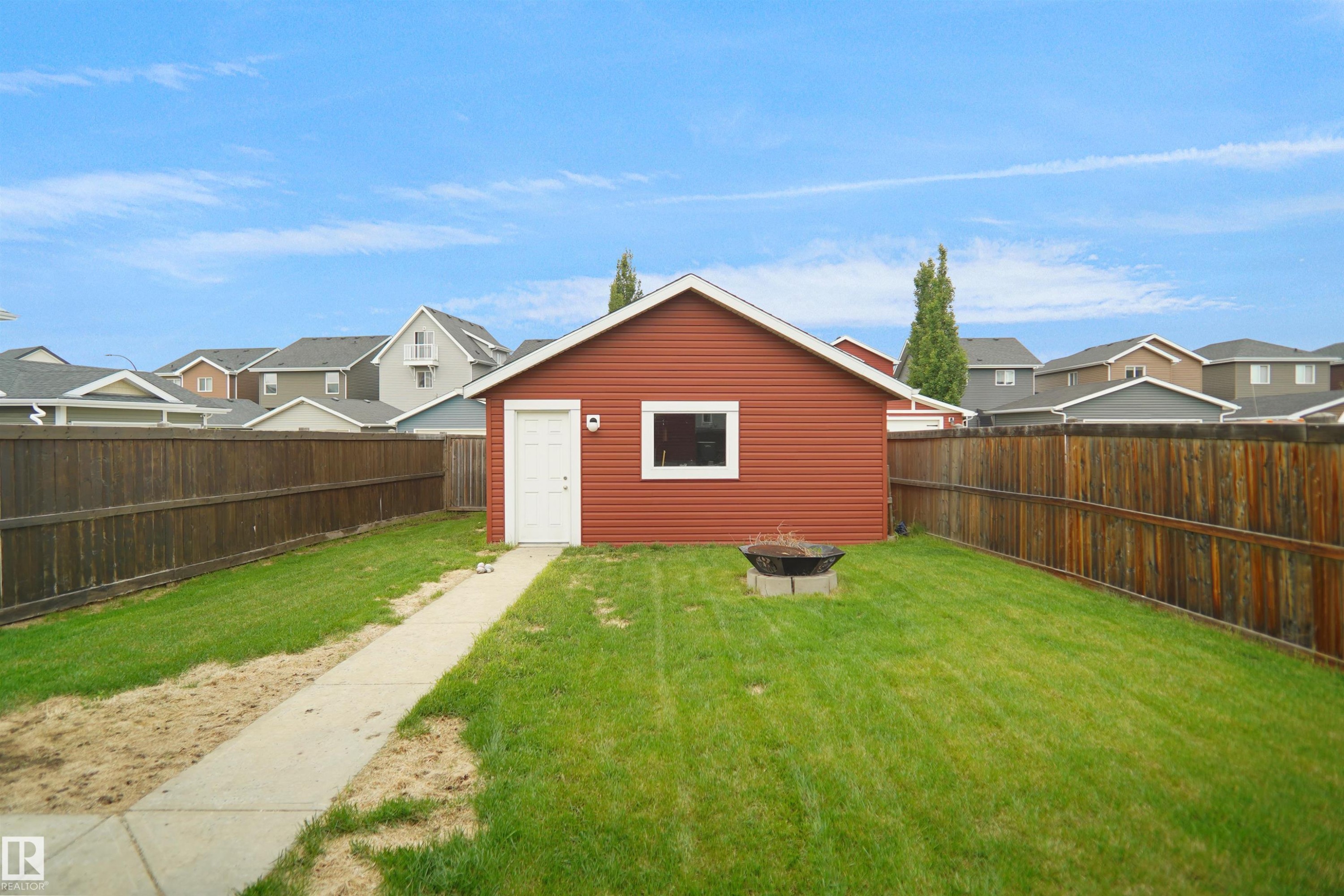 Detached double Garage with white entry door and window, featuring a concrete pathway, a fire pit, and a surrounding wooden fence - 292 Simmonds Way, Leduc, AB