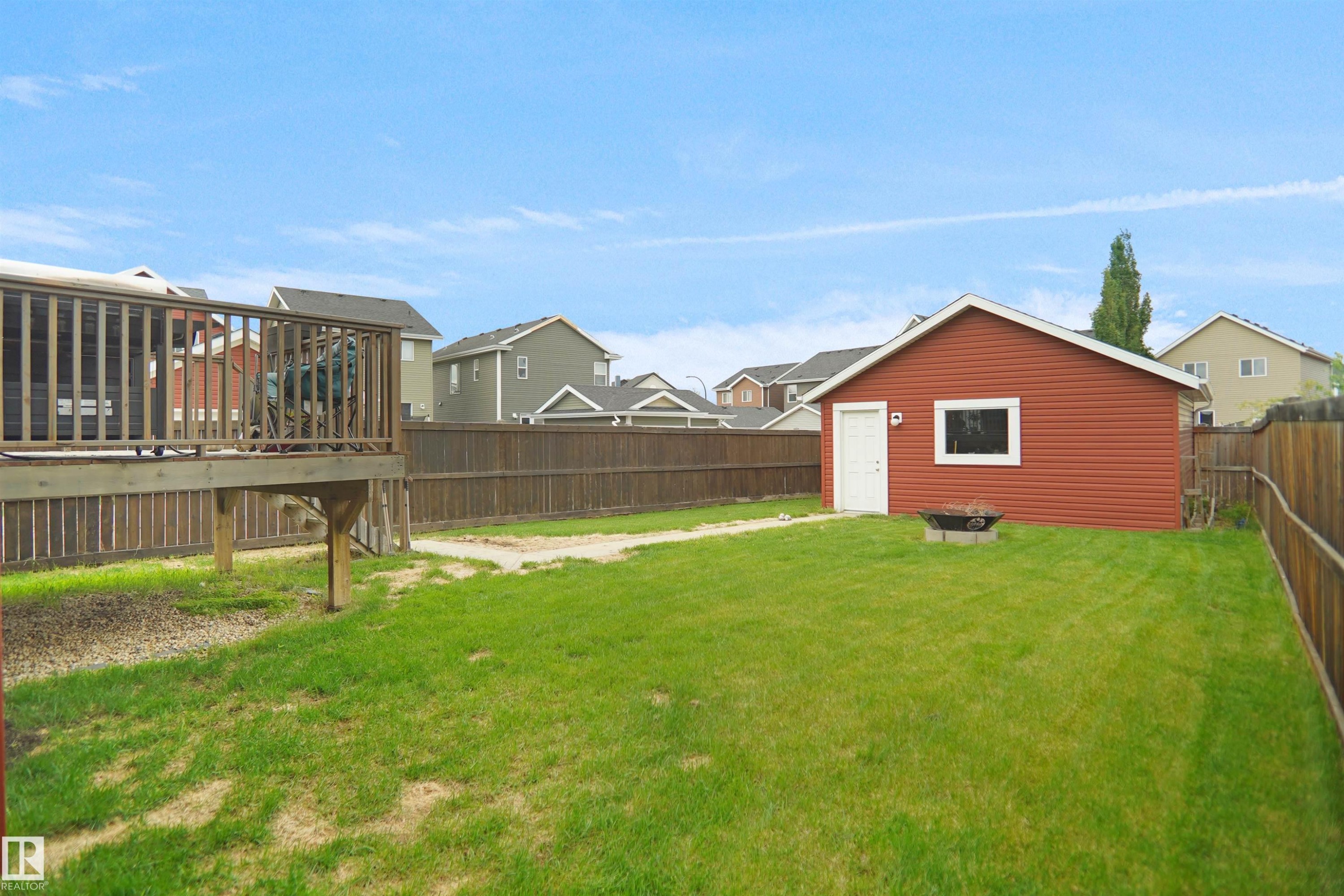 Expansive backyard featuring a manicured lawn, a detached red utility building with a white door and window, a wood privacy fence, and an elevated wood-plank deck with railings - 292 Simmonds Way, Leduc, AB