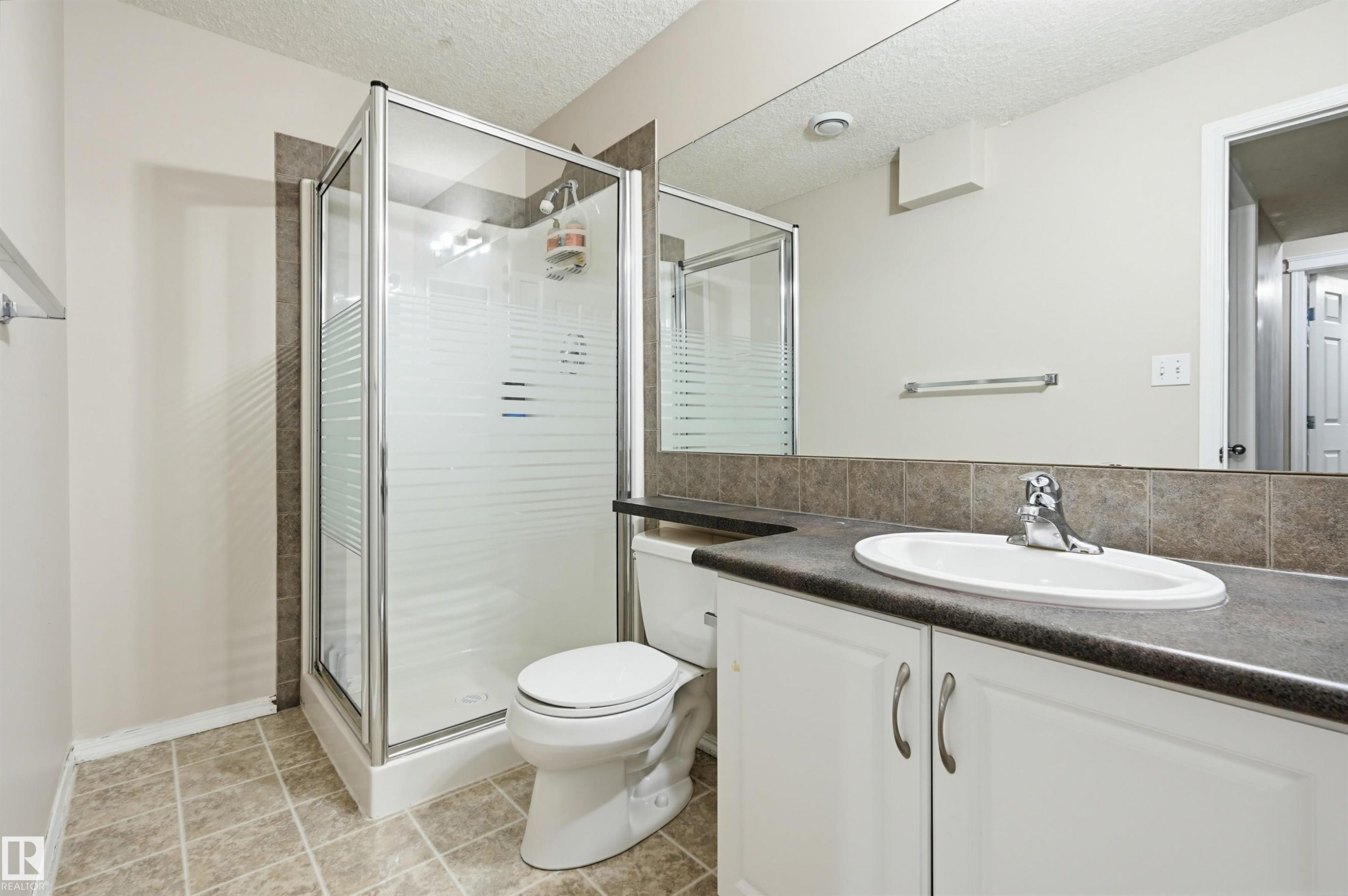 Bathroom featuring a glass-enclosed shower, white vanity cabinetry, a dark countertop, a rectangular mirror, and neutral-toned floor tiling - 1804 121 Street, Edmonton, AB