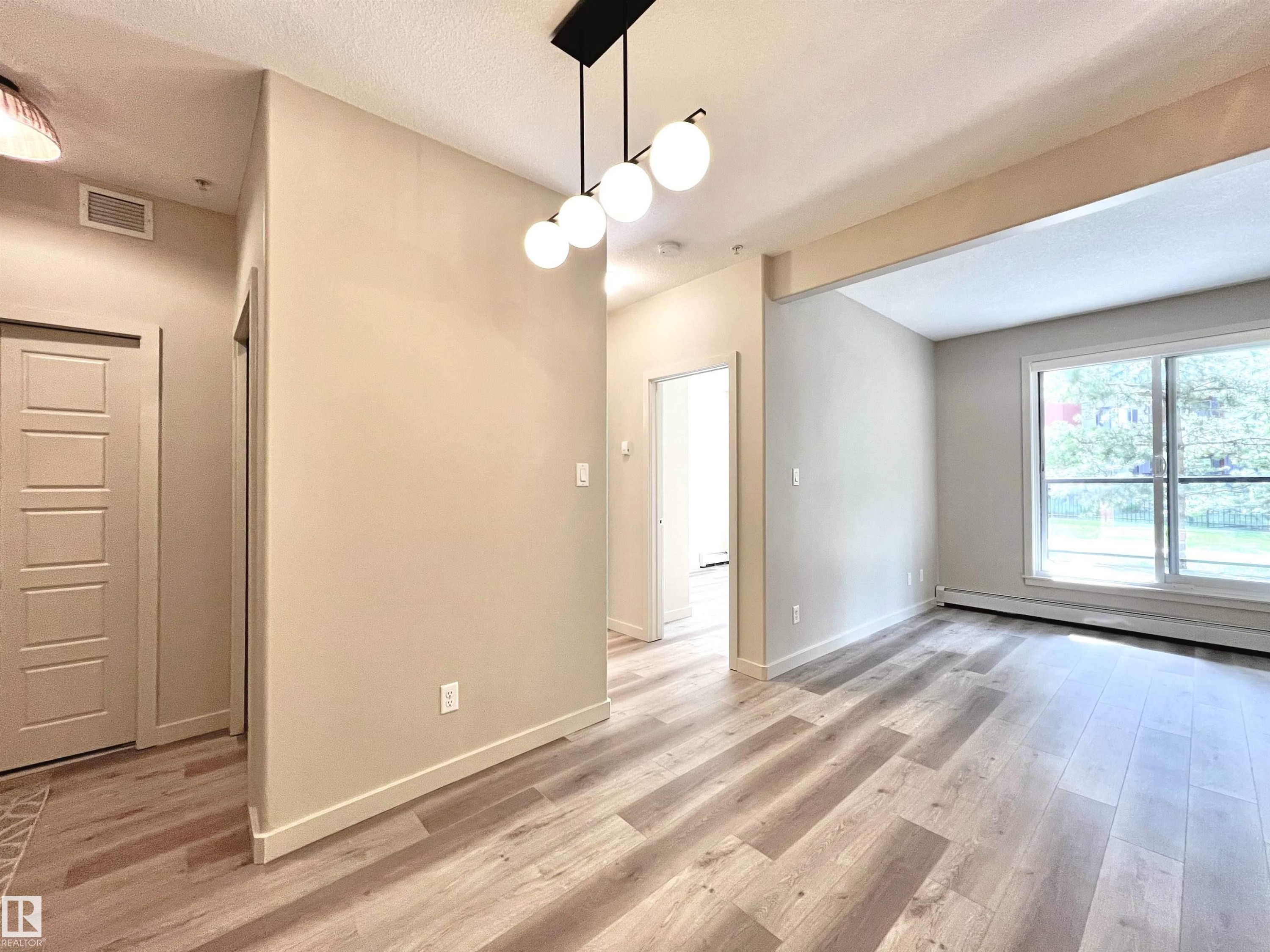Entryway and living area featuring wood-finish flooring, a modern linear chandelier with spherical lights, a panel door, and large sliding windows - 140 308 Ambleside Link Link, Edmonton, AB
