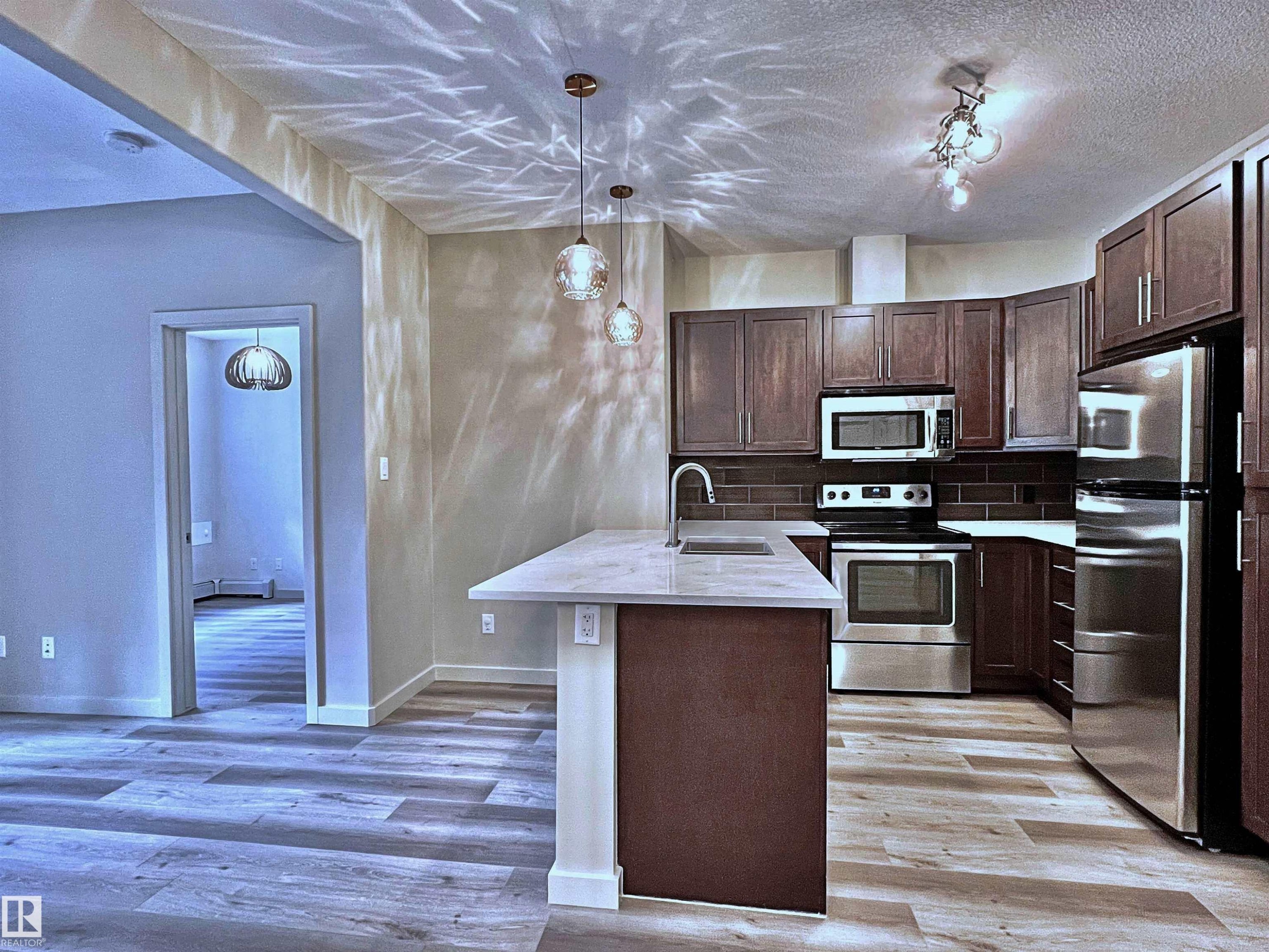 Modern kitchen featuring stainless steel appliances, dark wood-finish cabinetry, a large center island with a sink and pendant lighting, a subway tile backsplash, and light wood-finish flooring - 140 308 Ambleside Link Link, Edmonton, AB
