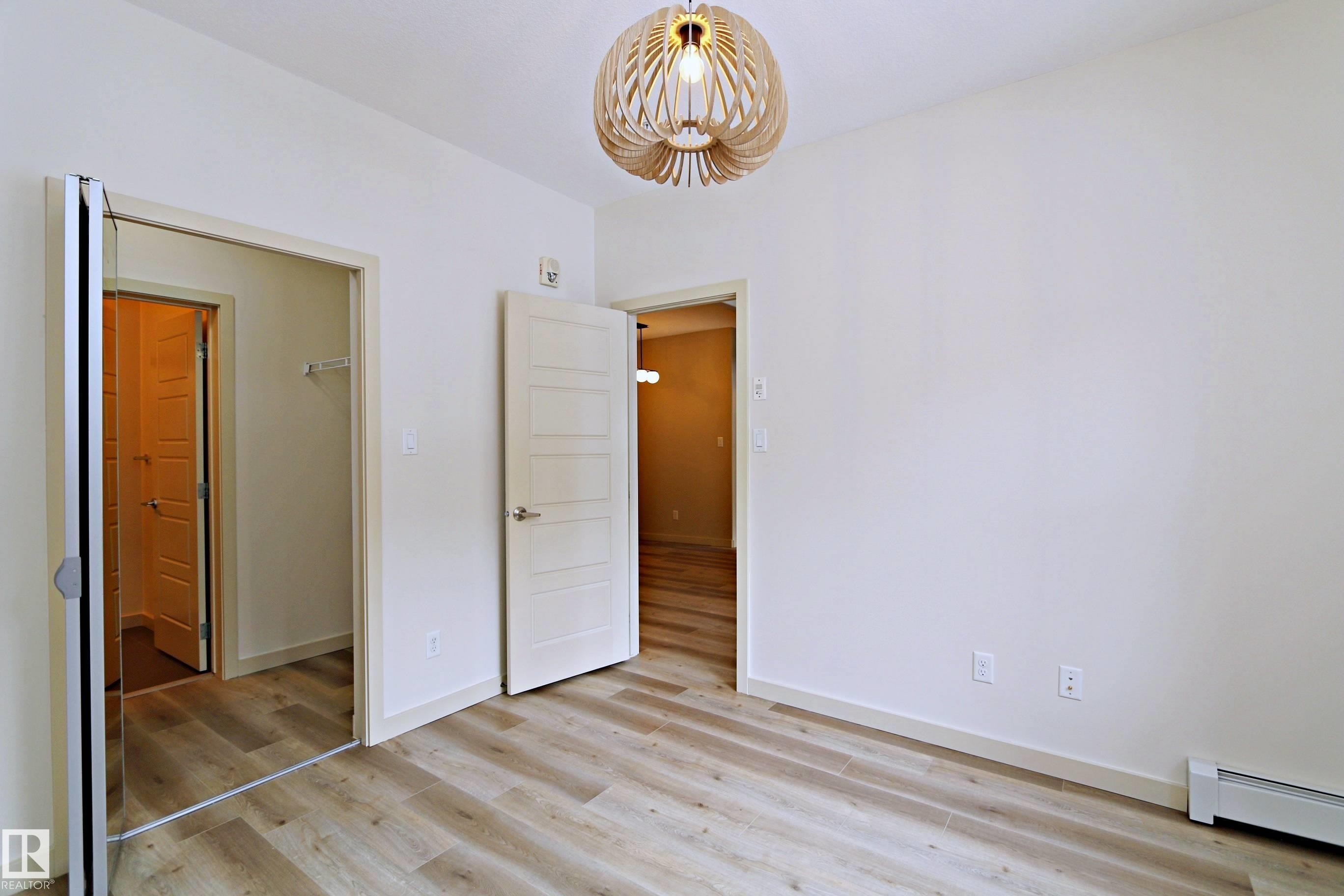 Spacious room featuring wood-finish flooring, a modern pendant light fixture, and a white interior door with horizontal paneling - 140 308 Ambleside Link Link, Edmonton, AB