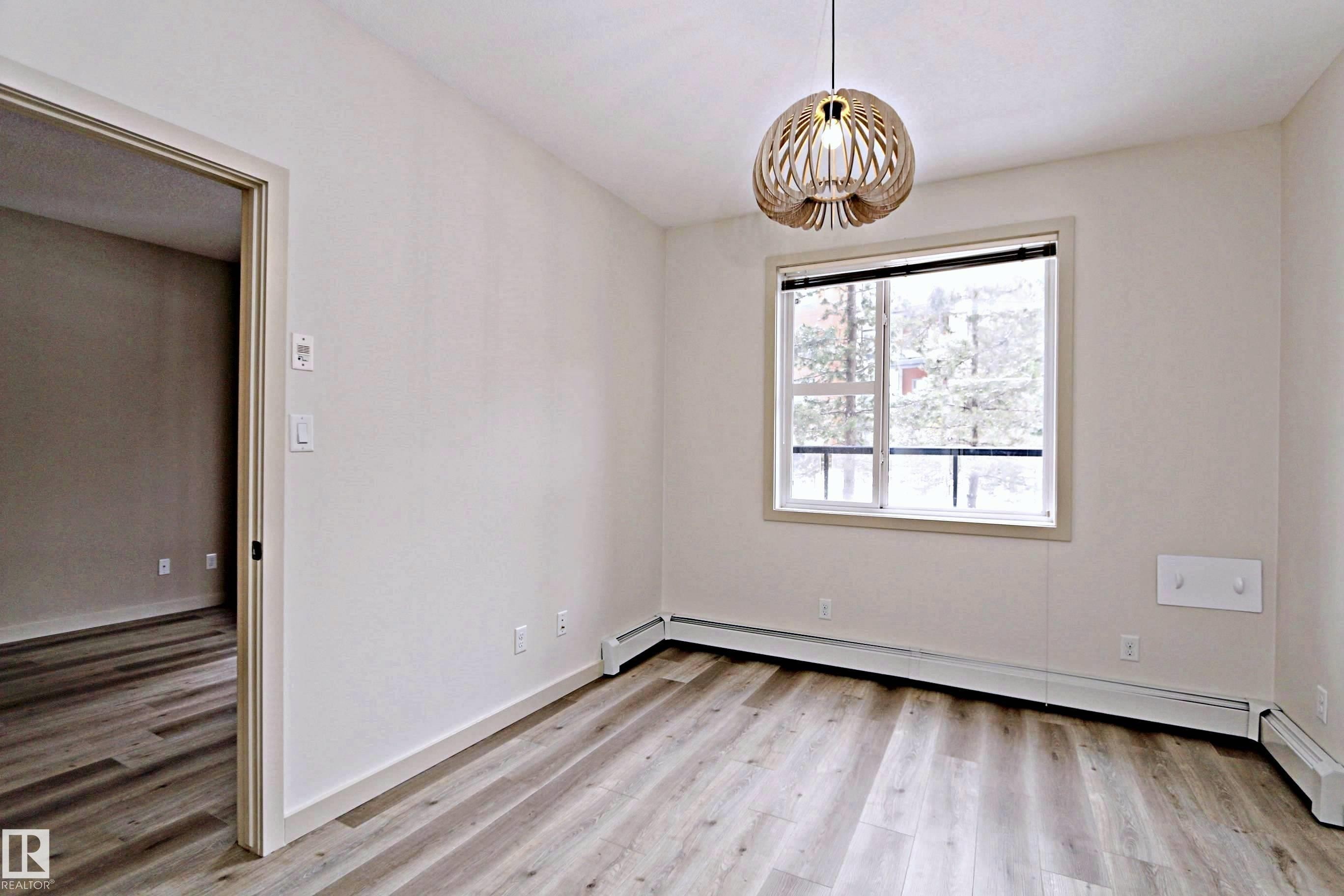 Bright room featuring a contemporary wood-slat pendant light, wood-finish flooring, a large window, and baseboard heating - 140 308 Ambleside Link Link, Edmonton, AB