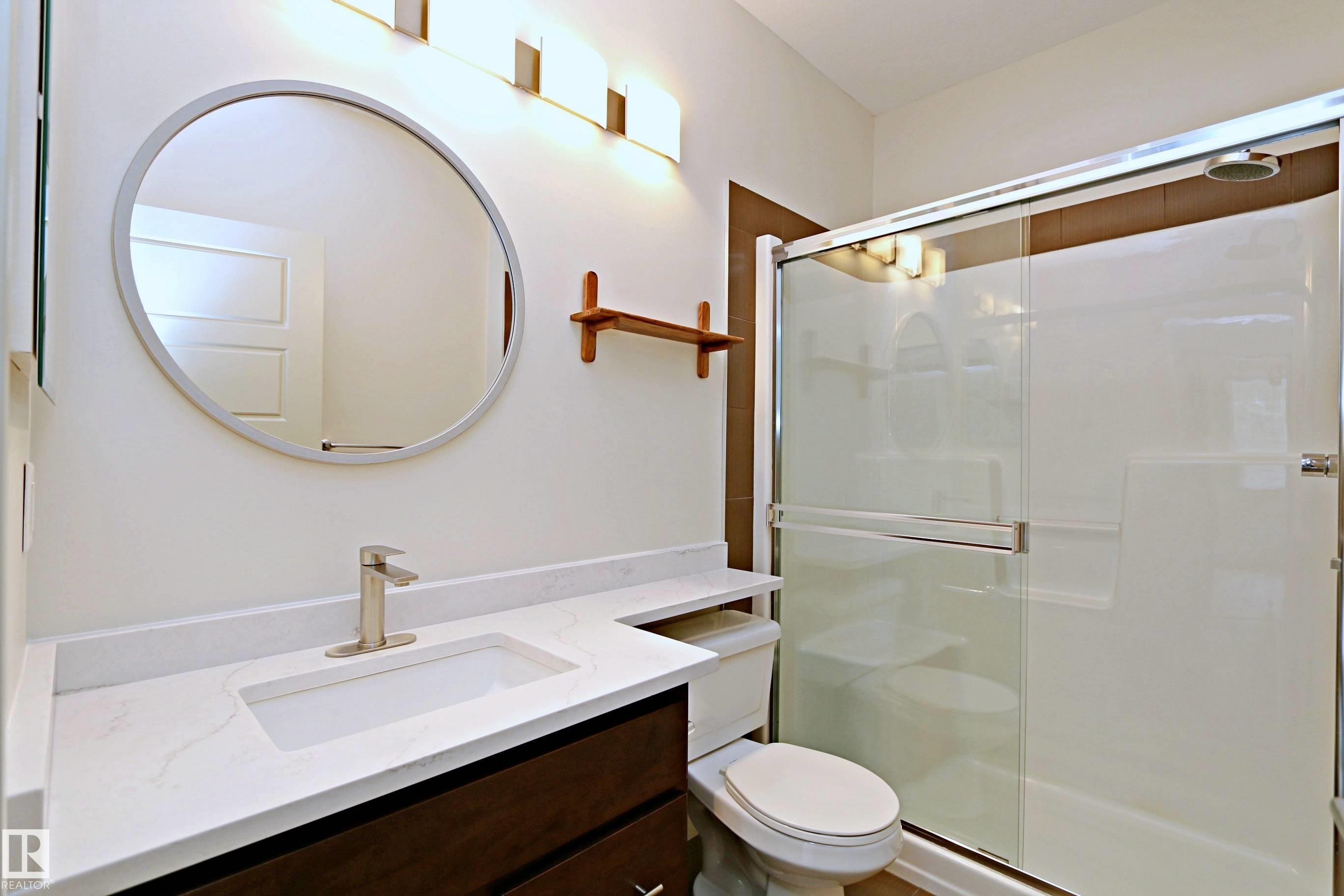 Bathroom featuring a dark wood-finish vanity with an integrated white sink, a white countertop, and a circular mirror - 140 308 Ambleside Link Link, Edmonton, AB