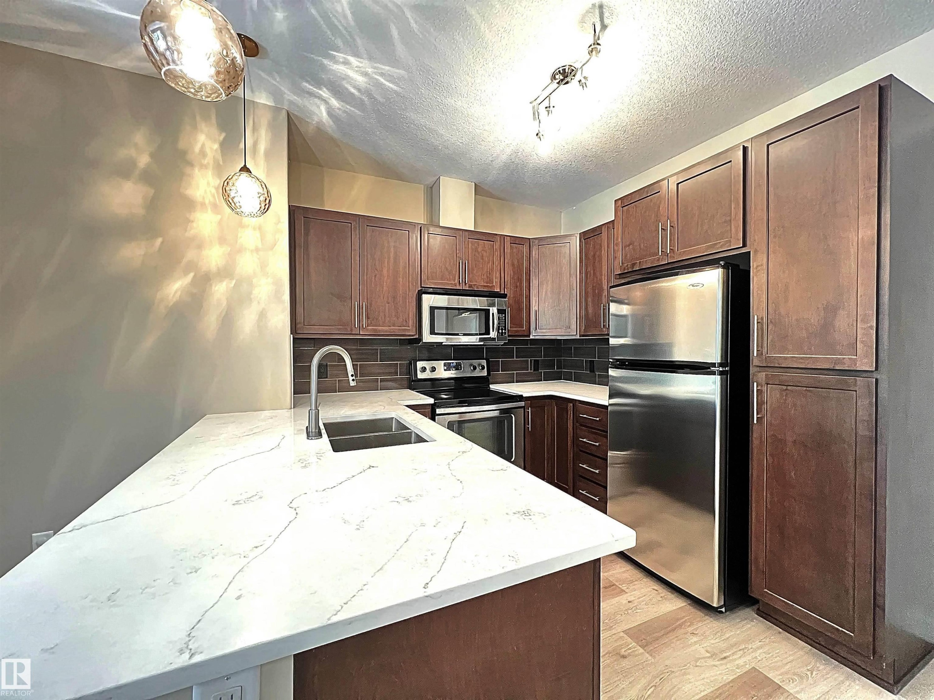 Modern kitchen featuring dark wood cabinetry, stainless steel appliances, a white stone-finish countertop with an undermount sink, and a dark subway tile backsplash - 140 308 Ambleside Link Link, Edmonton, AB