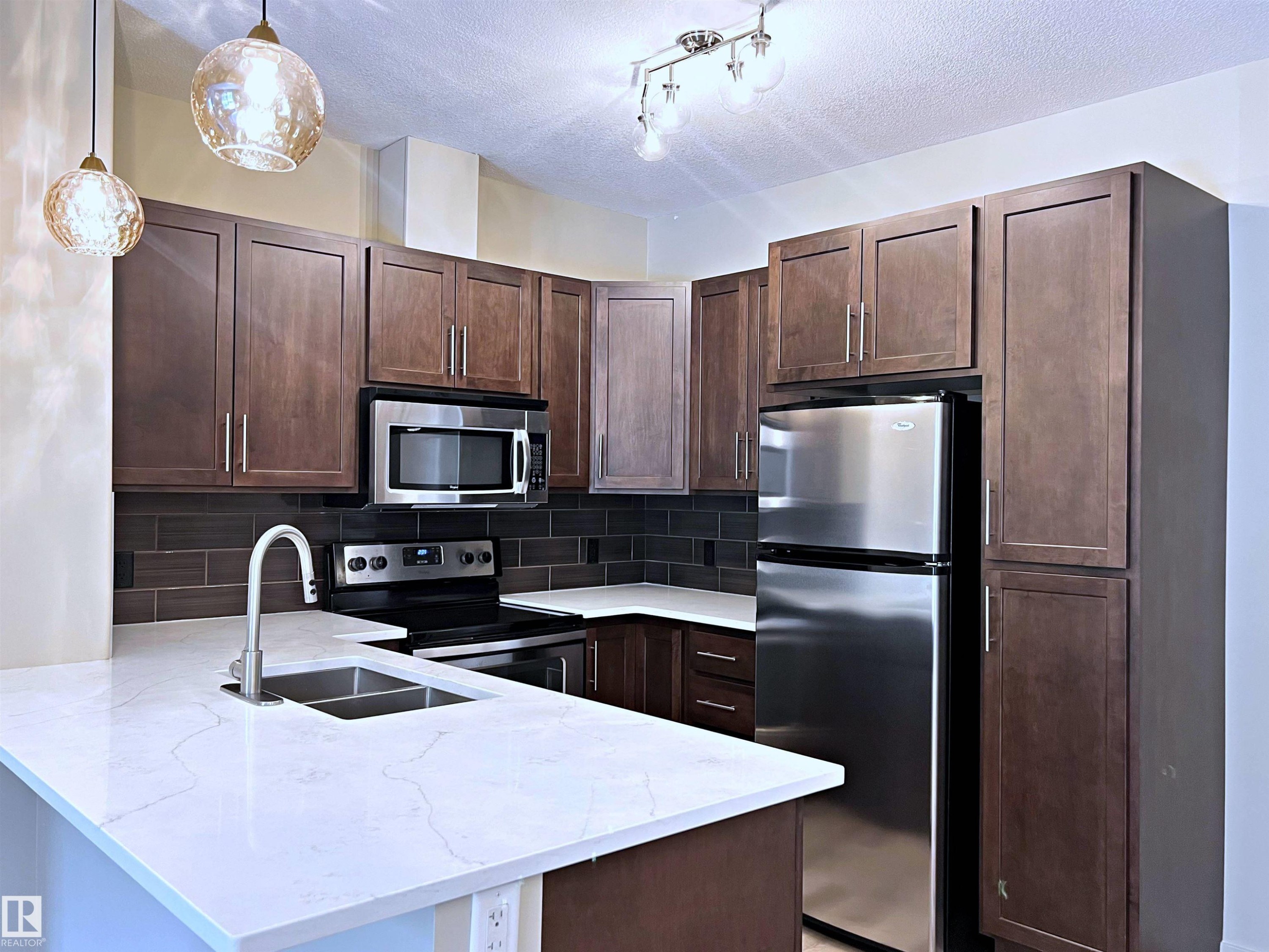 Kitchen featuring dark wood cabinetry, stainless steel appliances, a white stone-finish countertop, a dark tile backsplash, and decorative pendant lighting - 140 308 Ambleside Link Link, Edmonton, AB