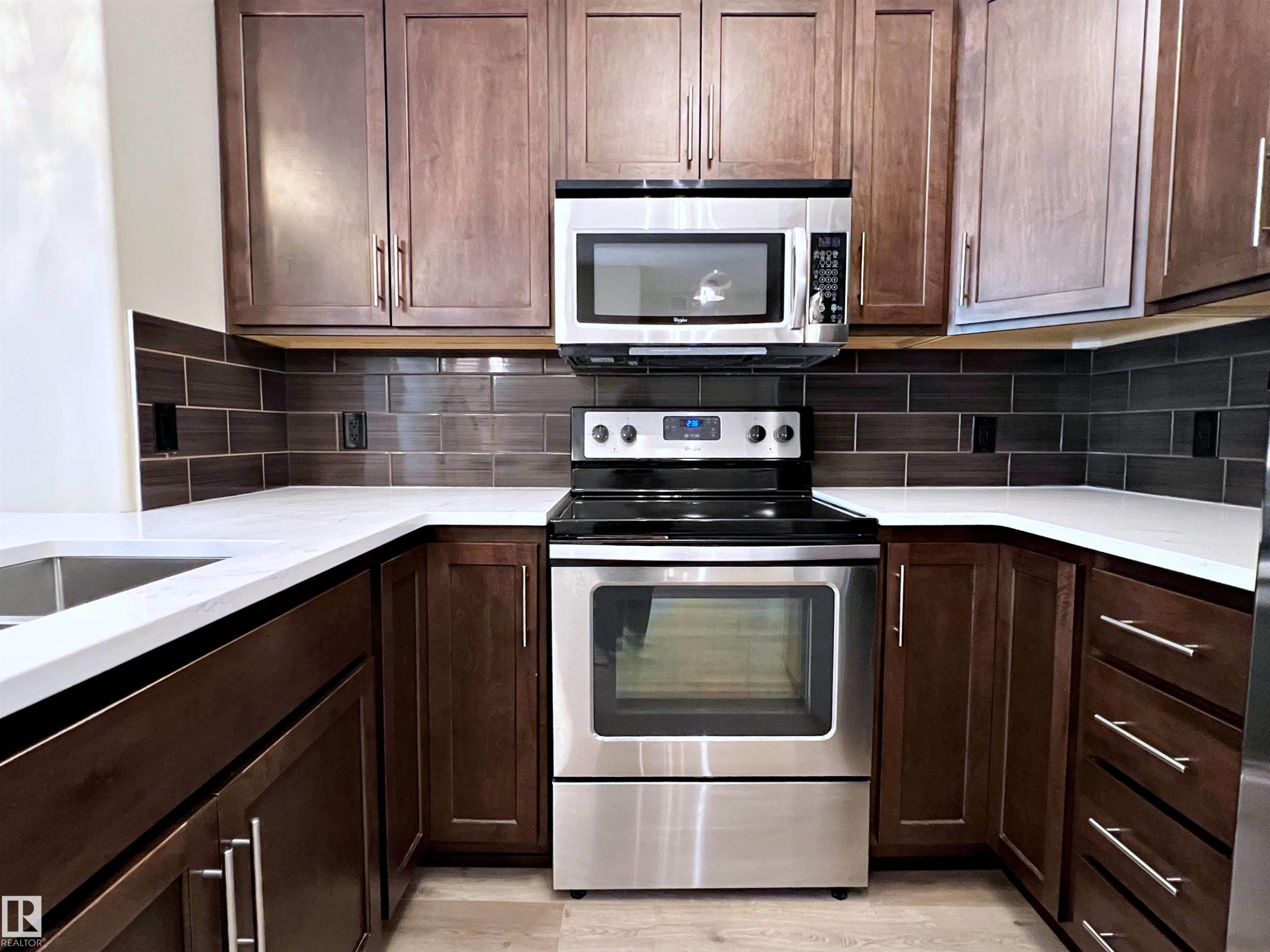 U-shaped kitchen featuring dark wood cabinetry with brushed nickel hardware, white countertops, and a dark subway tile backsplash - 140 308 Ambleside Link Link, Edmonton, AB