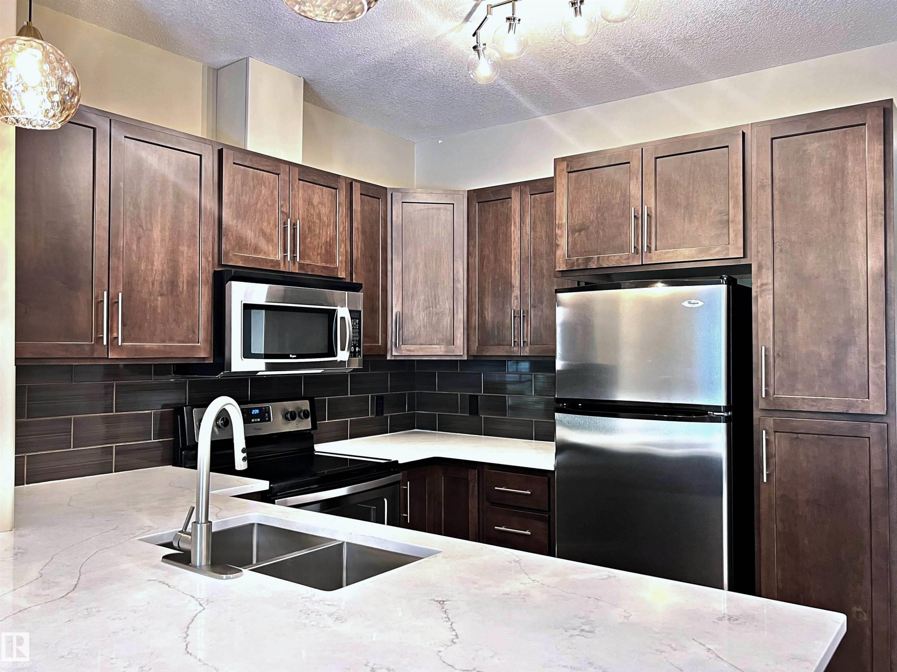 Modern kitchen featuring dark wood cabinetry, white stone-finish countertops, stainless steel appliances, a subway tile backsplash, and a double basin sink with a gooseneck faucet - 140 308 Ambleside Link Link, Edmonton, AB