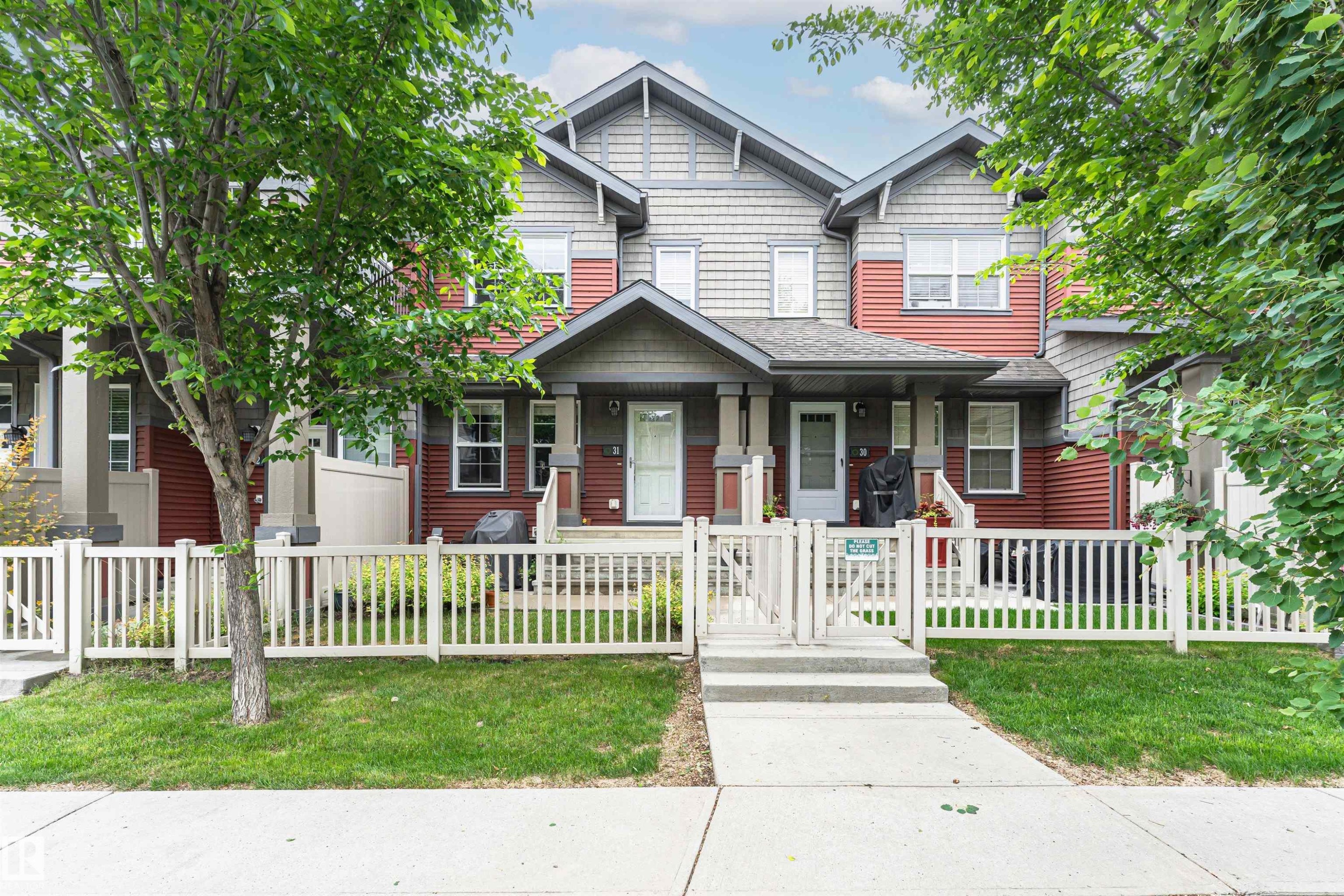 Multi-level townhouse exterior featuring vertical and horizontal siding, a fenced front yard with grass, mature trees, a concrete walkway, and a covered porch entry - 31 4029 Orchards Drive, Edmonton, AB