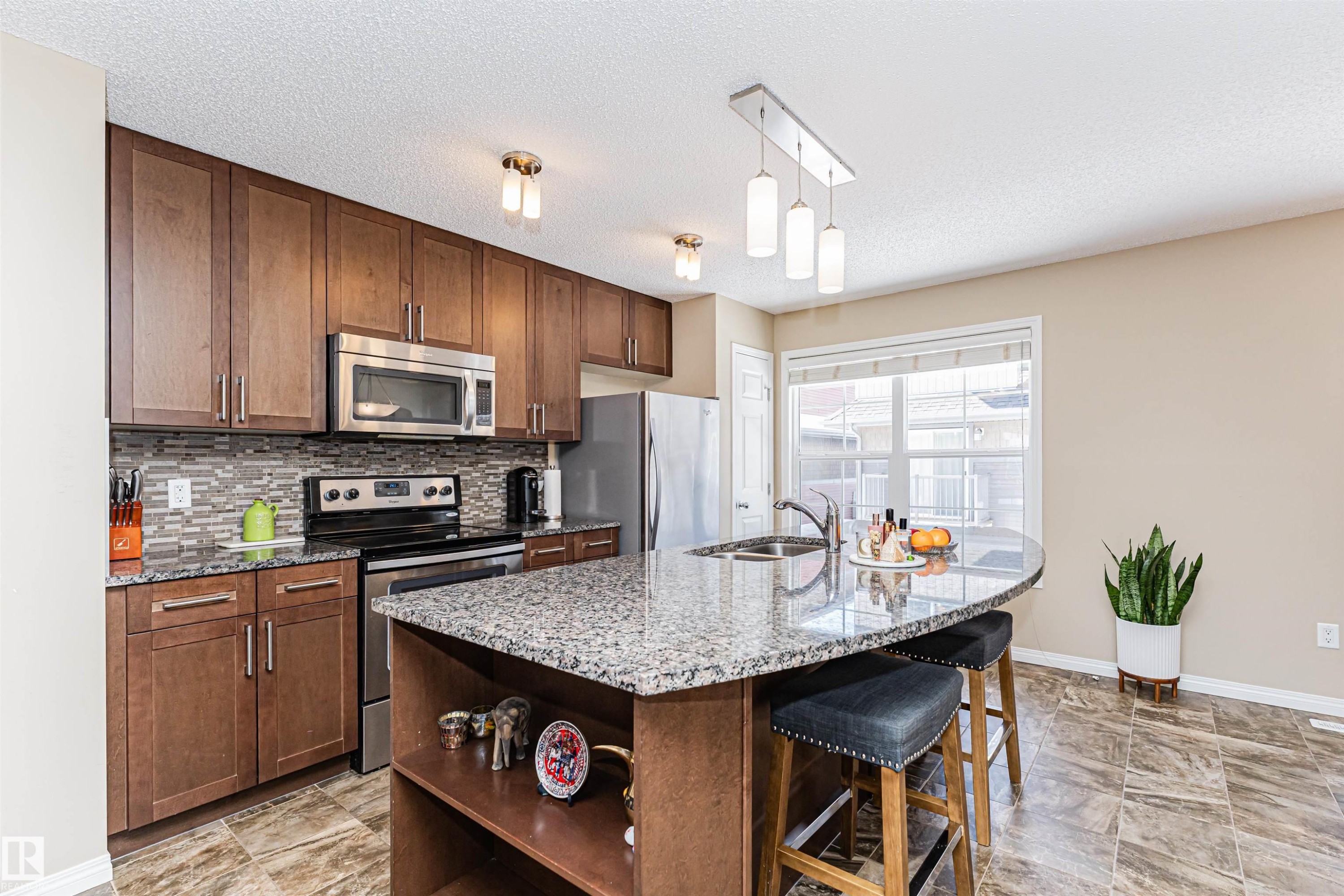Kitchen featuring a granite-topped island with breakfast bar seating, stainless steel appliances, wood-finish cabinetry, a tiled backsplash, and recessed lighting - 31 4029 Orchards Drive, Edmonton, AB