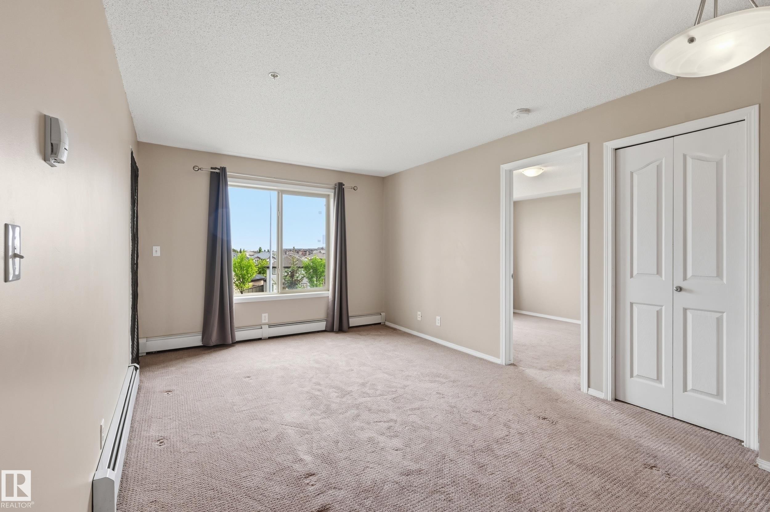 Carpeted living area featuring a large window, neutral wall tones, white baseboards, and a semi-flush ceiling light fixture - 304 11804 22 Avenue, Edmonton, AB