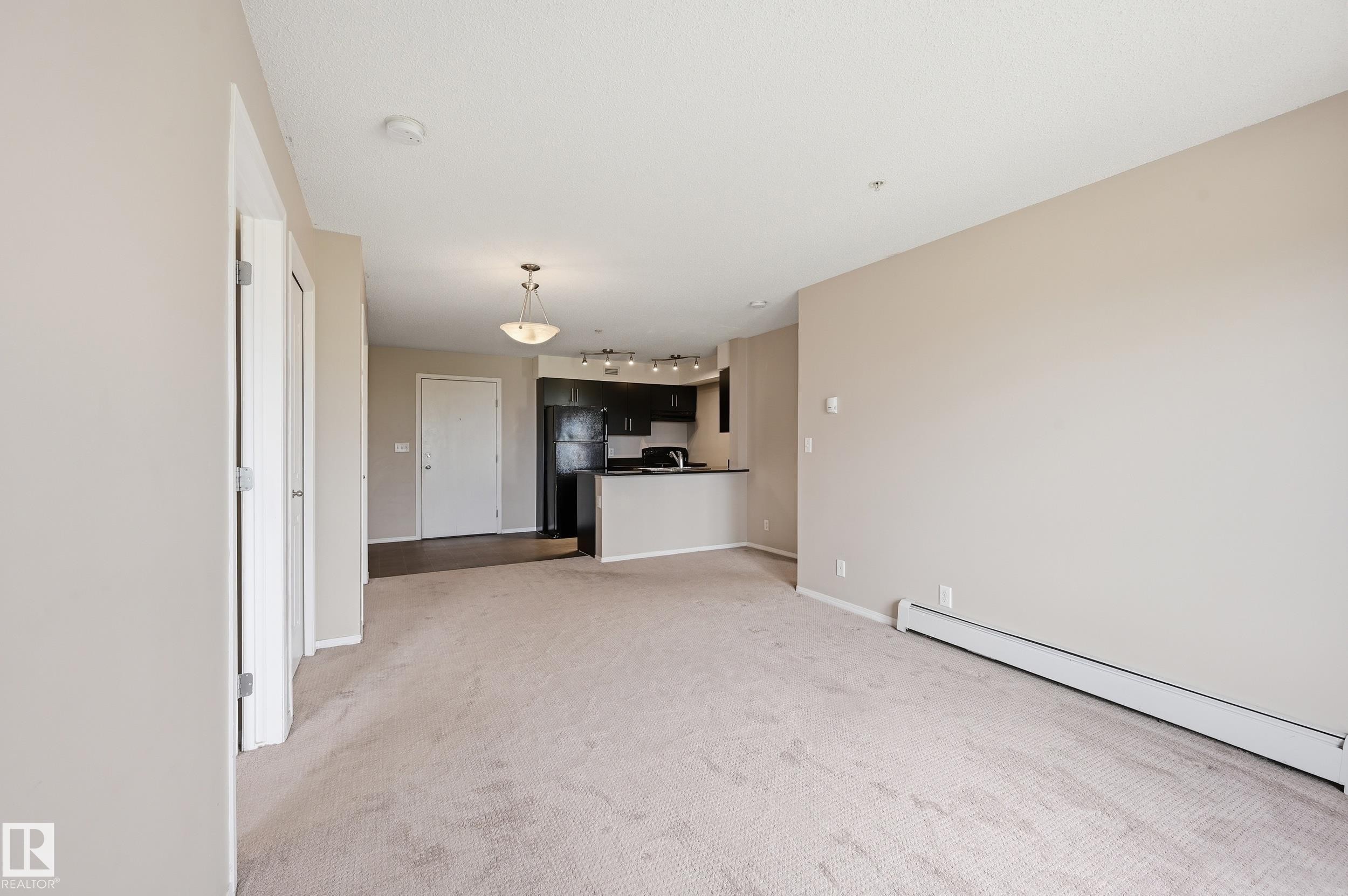 Open-concept living area with neutral carpeting, leading to a kitchen featuring dark cabinetry and black appliances - 304 11804 22 Avenue, Edmonton, AB