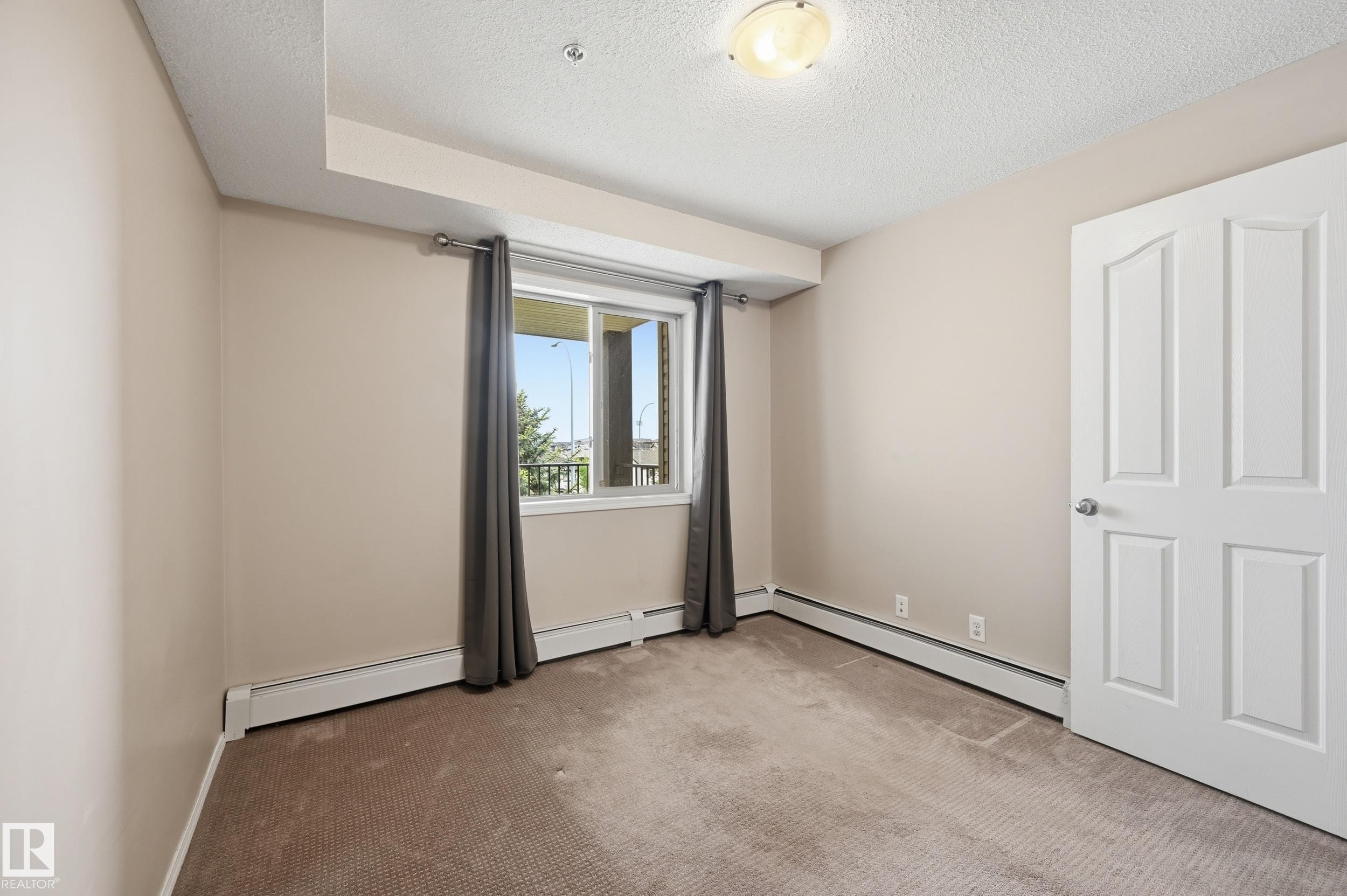 Carpeted room featuring a window with exterior scenery, a white paneled door, and baseboard heating - 304 11804 22 Avenue, Edmonton, AB