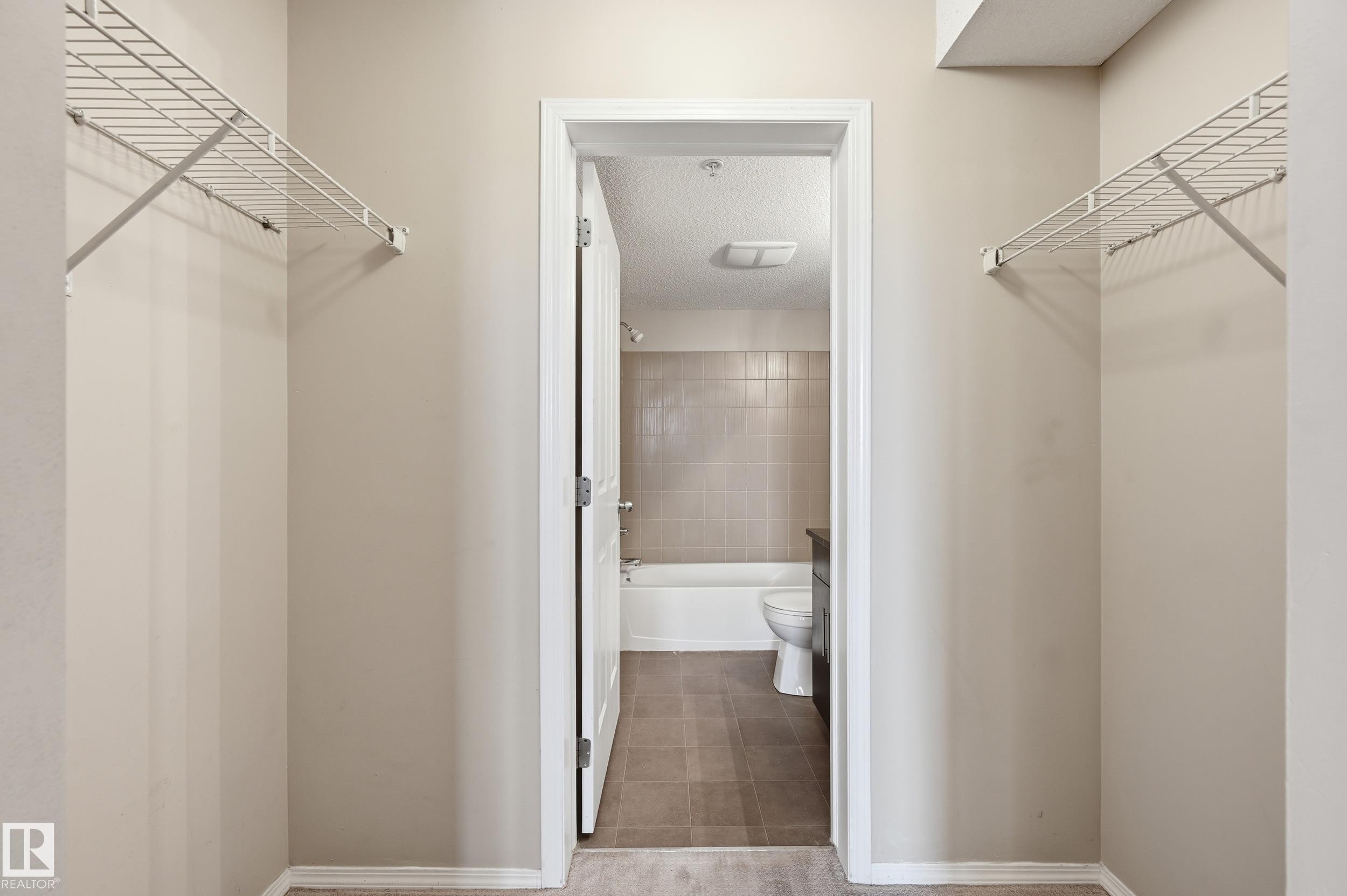 Alcove with light neutral wall paint and white wire shelving, opening to a bathroom with a white tub and tiled walls - 304 11804 22 Avenue, Edmonton, AB