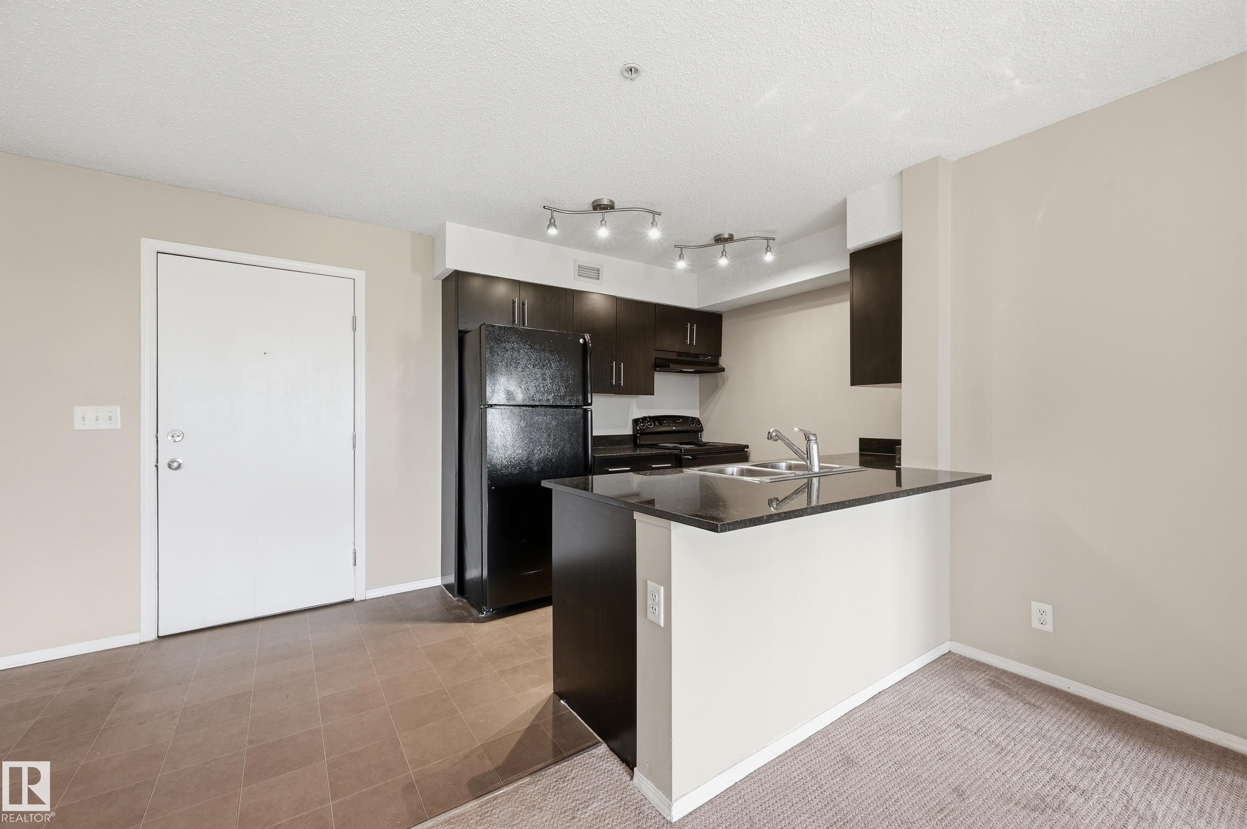 Kitchen featuring dark cabinetry, black appliances, a double basin stainless steel sink, and a breakfast bar with a dark countertop - 304 11804 22 Avenue, Edmonton, AB