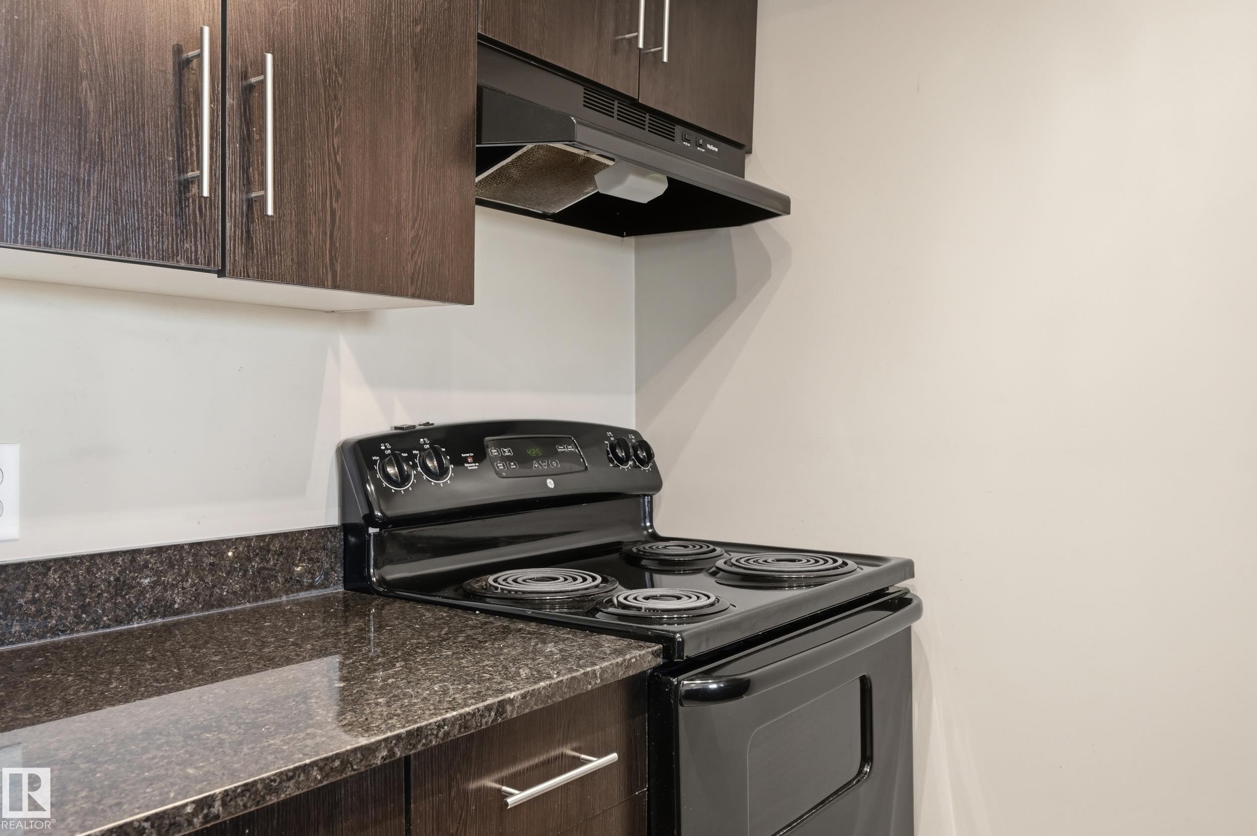 Kitchen featuring dark wood-finish cabinetry with silver hardware, speckled dark countertops, and a black electric range with an overhead vent - 304 11804 22 Avenue, Edmonton, AB