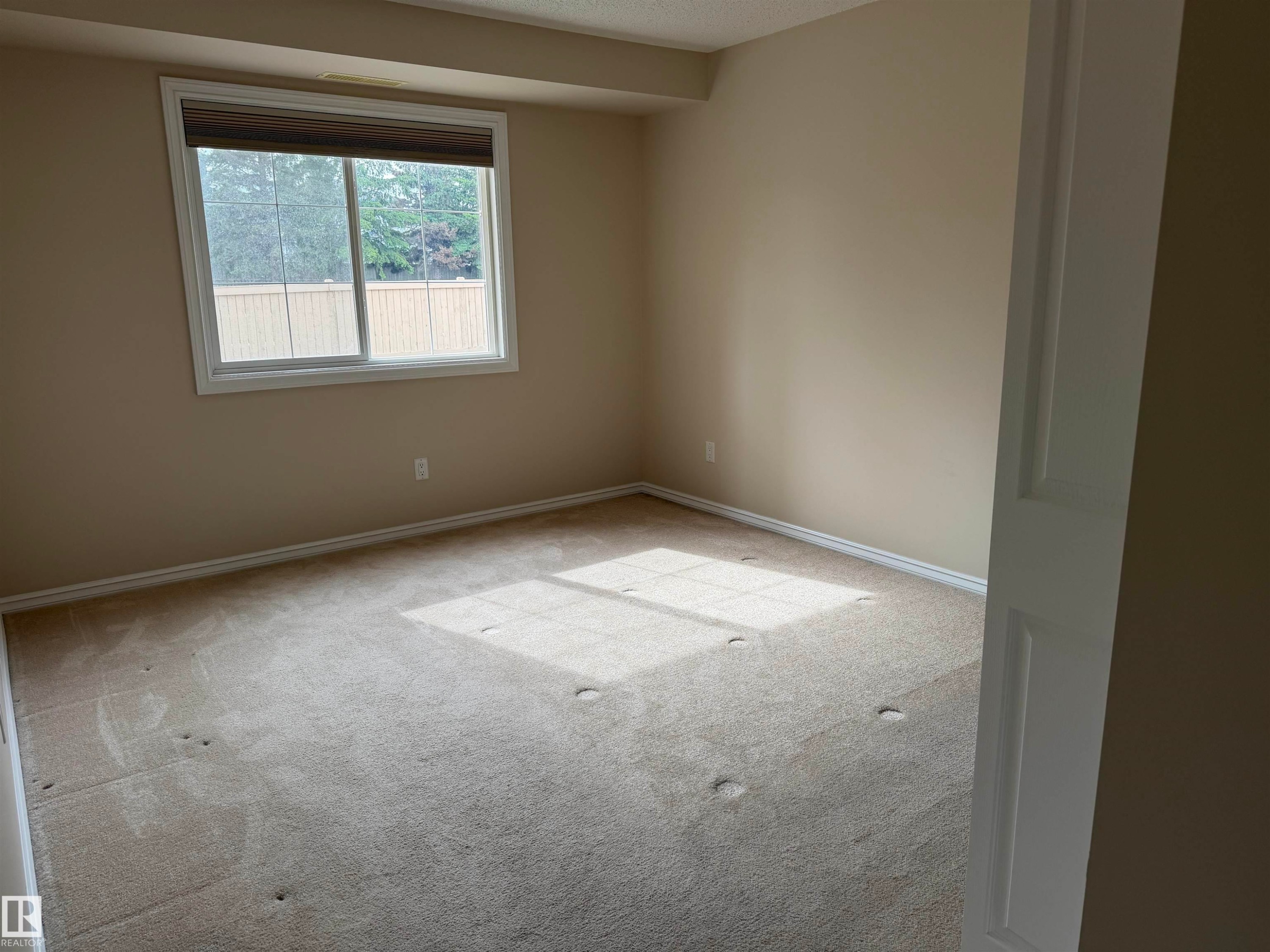 Carpeted interior room featuring a large window with white trim, neutral wall color, and white baseboards - 118 7801 Golf Course Road, Stony Plain, AB