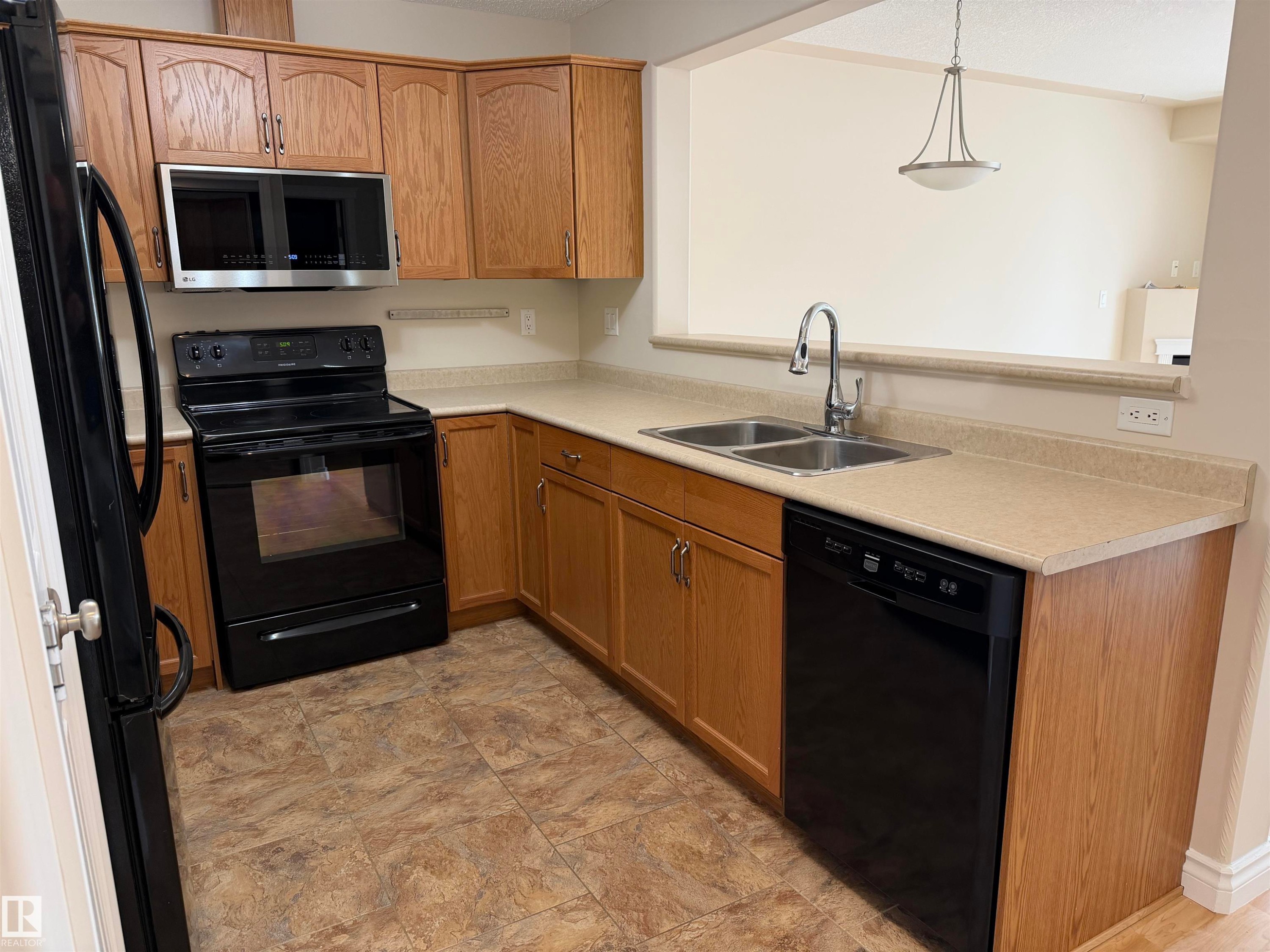 L-shaped kitchen featuring wood cabinetry, laminate countertops, a double basin stainless steel sink with a high-arc faucet, and black appliances including a range, microwave, and dishwasher - 118 7801 Golf Course Road, Stony Plain, AB