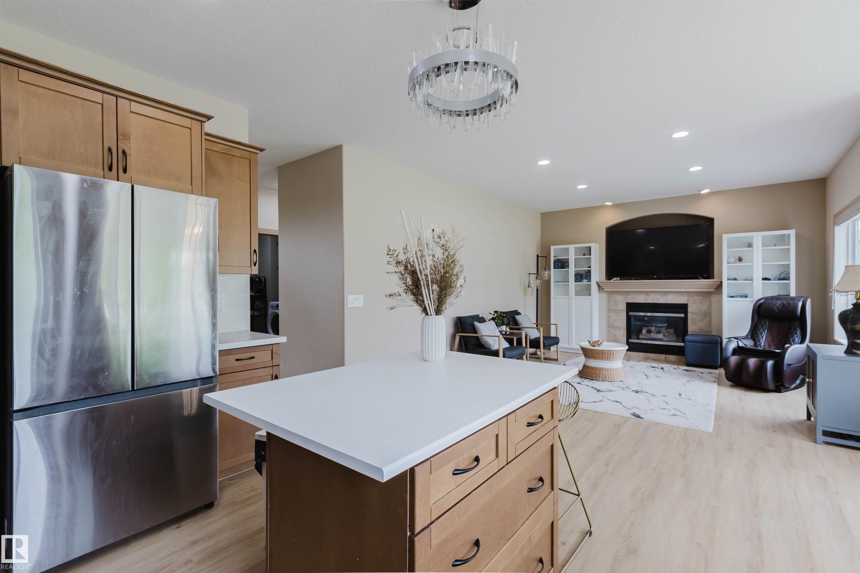 Kitchen island with white countertop and integrated storage, stainless steel French door refrigerator, wood-finish flooring, recessed lighting, and a fireplace with a mantel - 2236 Garnett Crest, Edmonton, AB