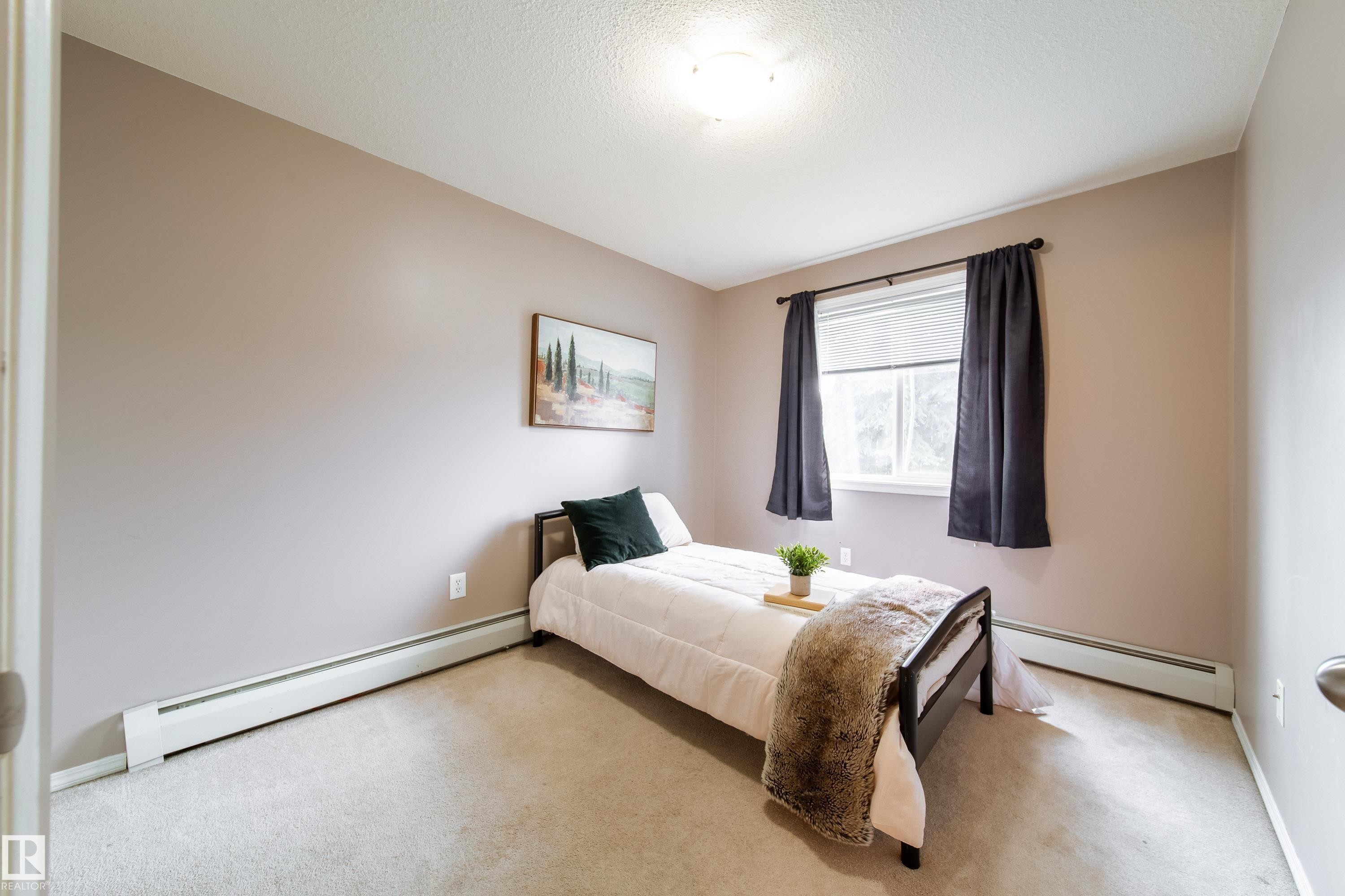 Carpeted bedroom featuring a single window with dark drapery, light-colored walls, and baseboard heating - 115 4309 33 Street, Stony Plain, AB