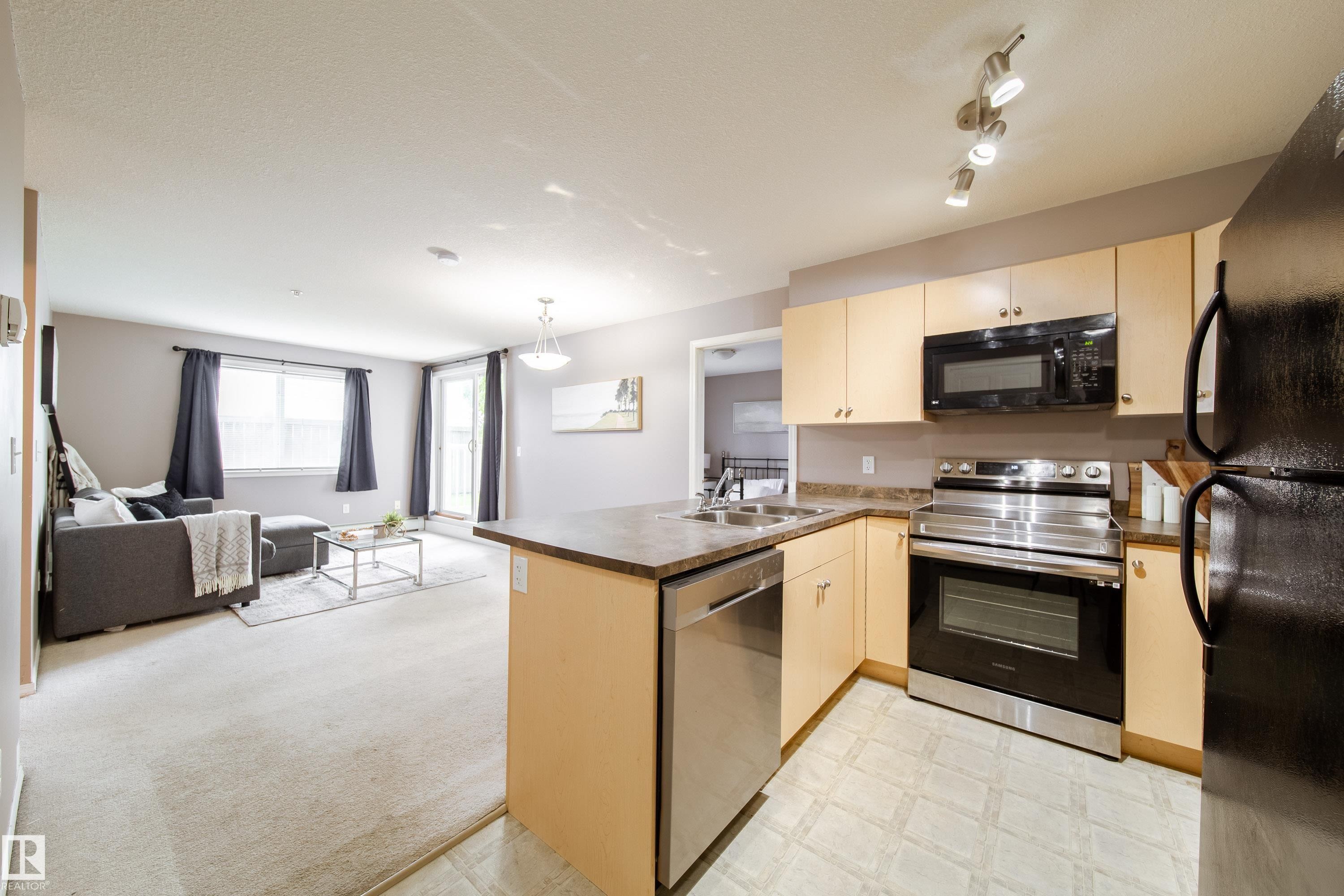 Open-concept living space featuring a kitchen with light wood-finish cabinetry and stainless steel appliances, a breakfast bar, and a living area with carpet flooring and a balcony access point - 115 4309 33 Street, Stony Plain, AB