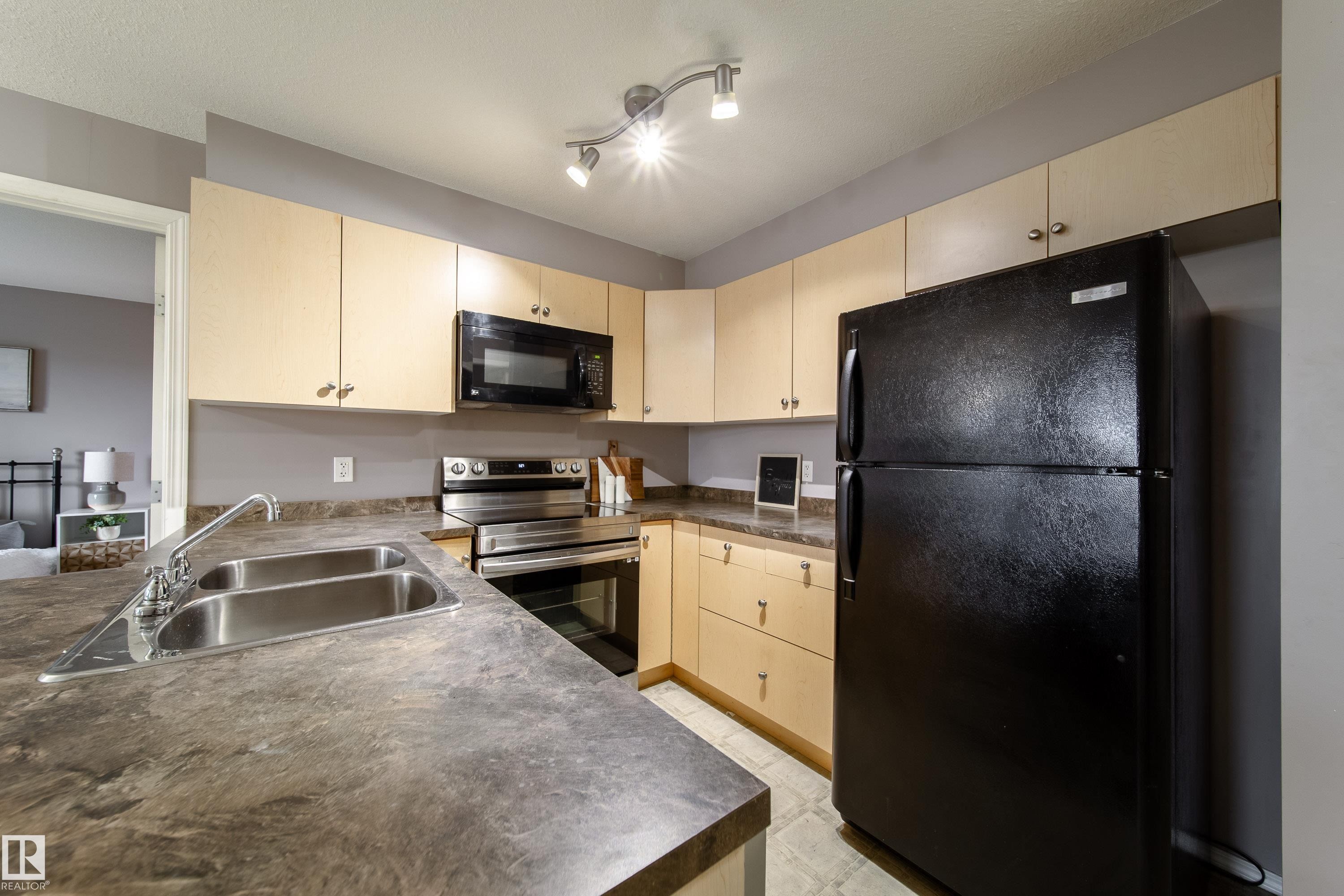 Kitchen featuring light wood-finish cabinetry, dark countertops, a double basin stainless steel sink, and a black refrigerator - 115 4309 33 Street, Stony Plain, AB