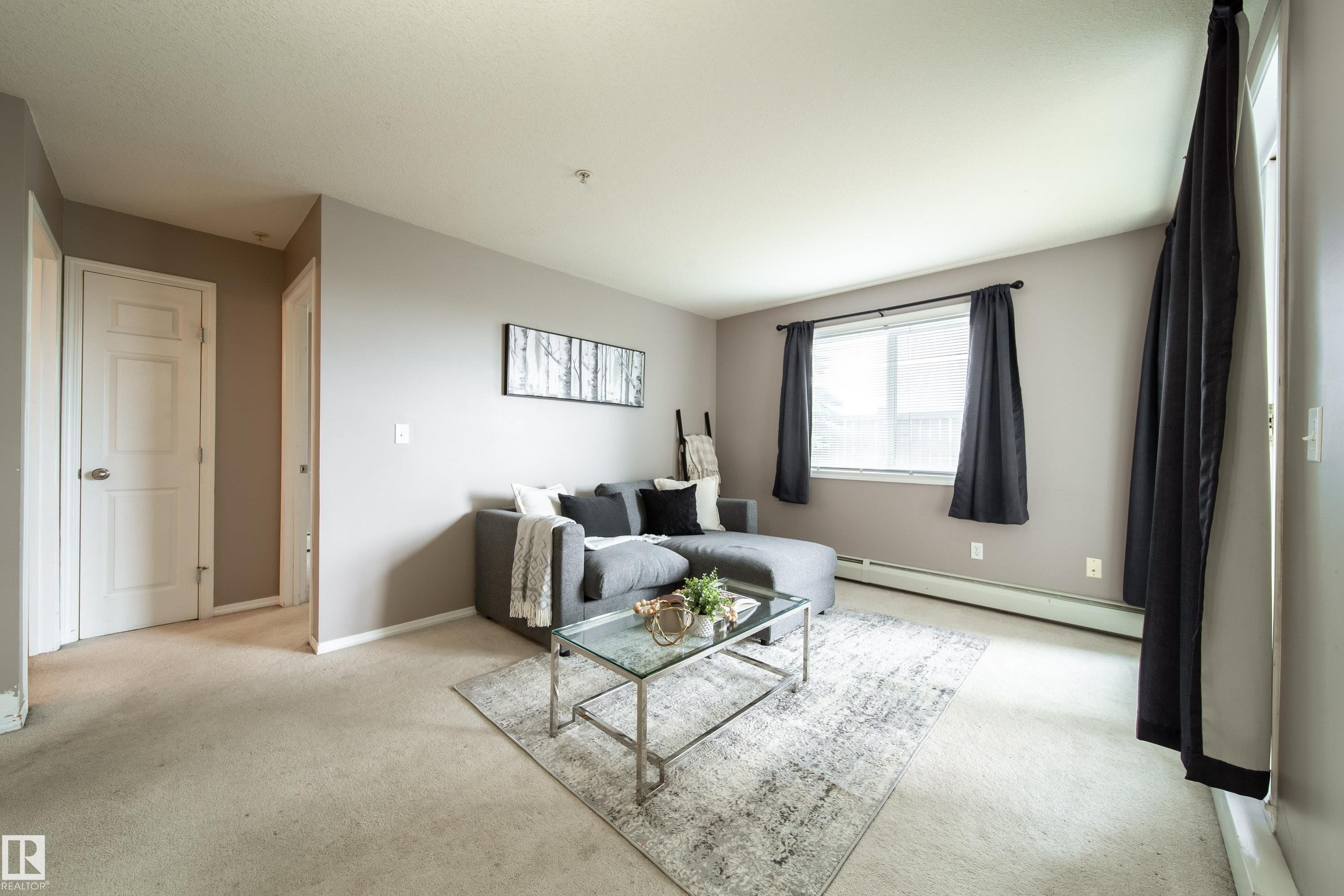 Living area featuring neutral wall tones, carpet flooring, baseboard heating, a white-framed window, and white interior doors - 115 4309 33 Street, Stony Plain, AB