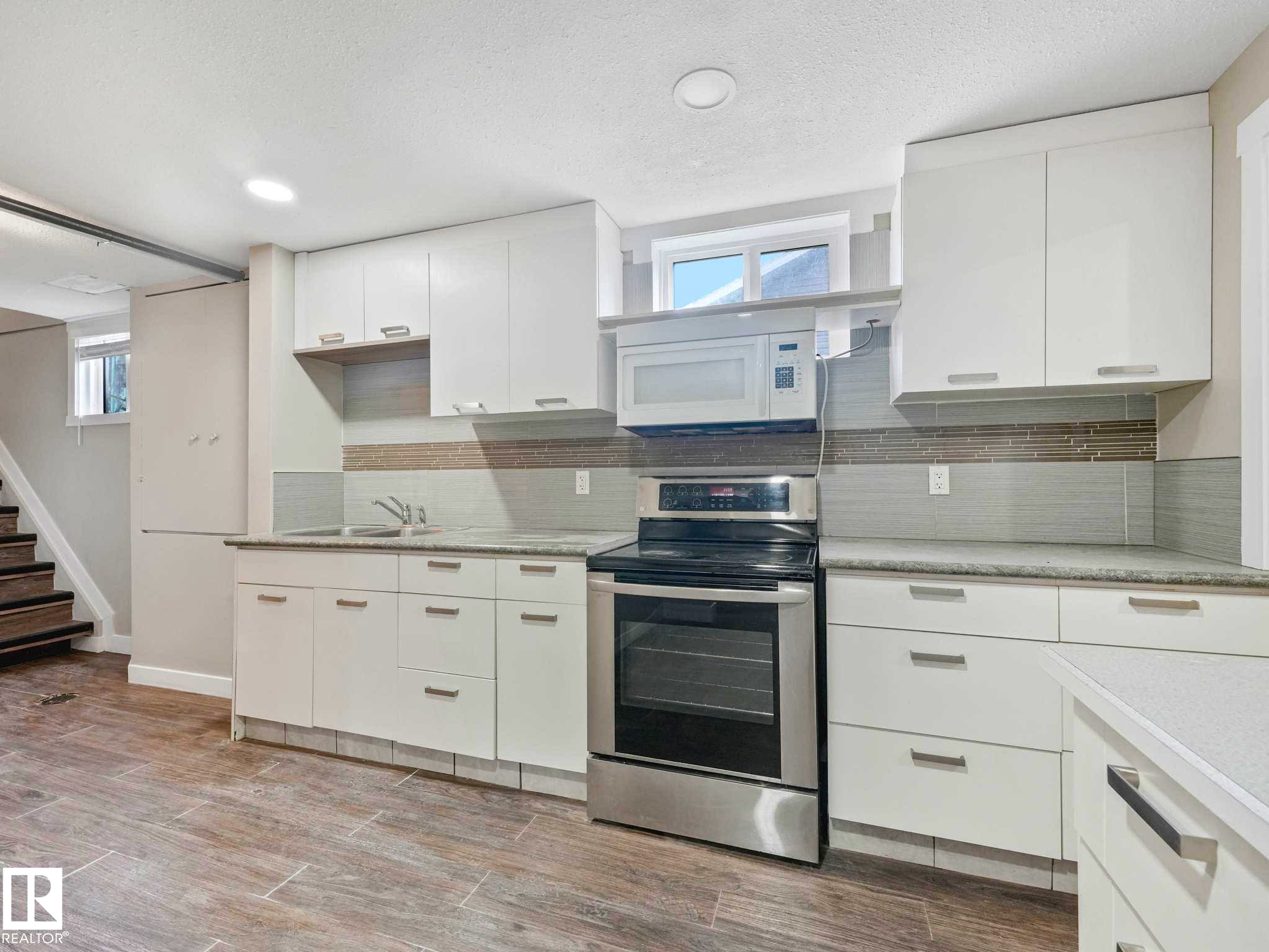 Contemporary kitchen featuring white cabinetry, stainless steel range and microwave, light-toned countertops, and a gray-toned tile backsplash - 12241 54 Street Nw, Edmonton, AB - Indoor Photo Showing Kitchen With Double Sink