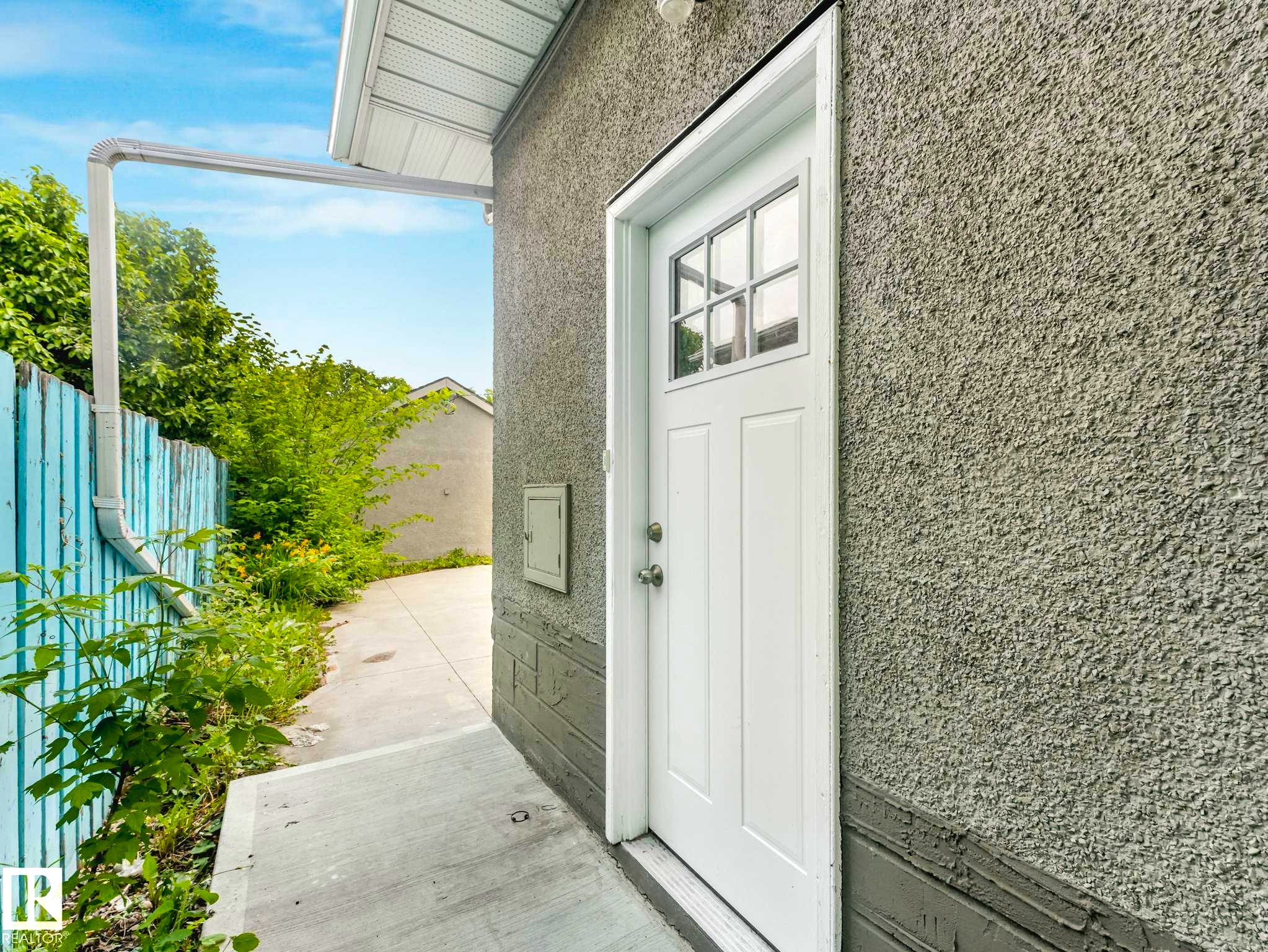 Side exterior entry featuring a white paneled door with windowpanes, stucco siding, and a concrete pathway - 12241 54 Street Nw, Edmonton, AB - Outdoor