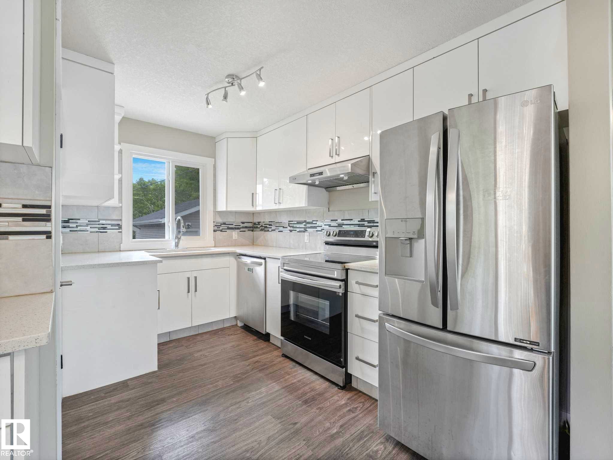 Modern kitchen featuring white cabinetry, stainless steel appliances, light-toned countertops, a mosaic tile backsplash, and wood-finish flooring - 12241 54 Street Nw, Edmonton, AB - Indoor Photo Showing Kitchen With Upgraded Kitchen